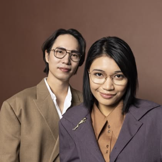 A man and woman wearing low-bridge Asian-fit glasses posing together in front of a brown background.