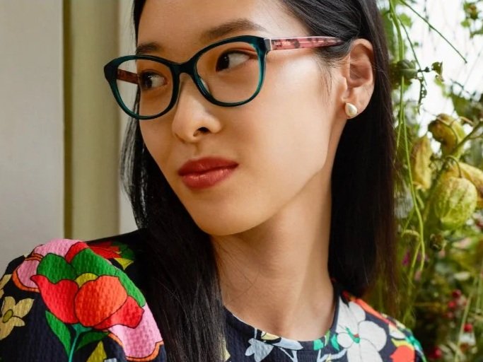 Close-up of a woman wearing colorful floral clothing and glasses, standing outdoors near green plants.