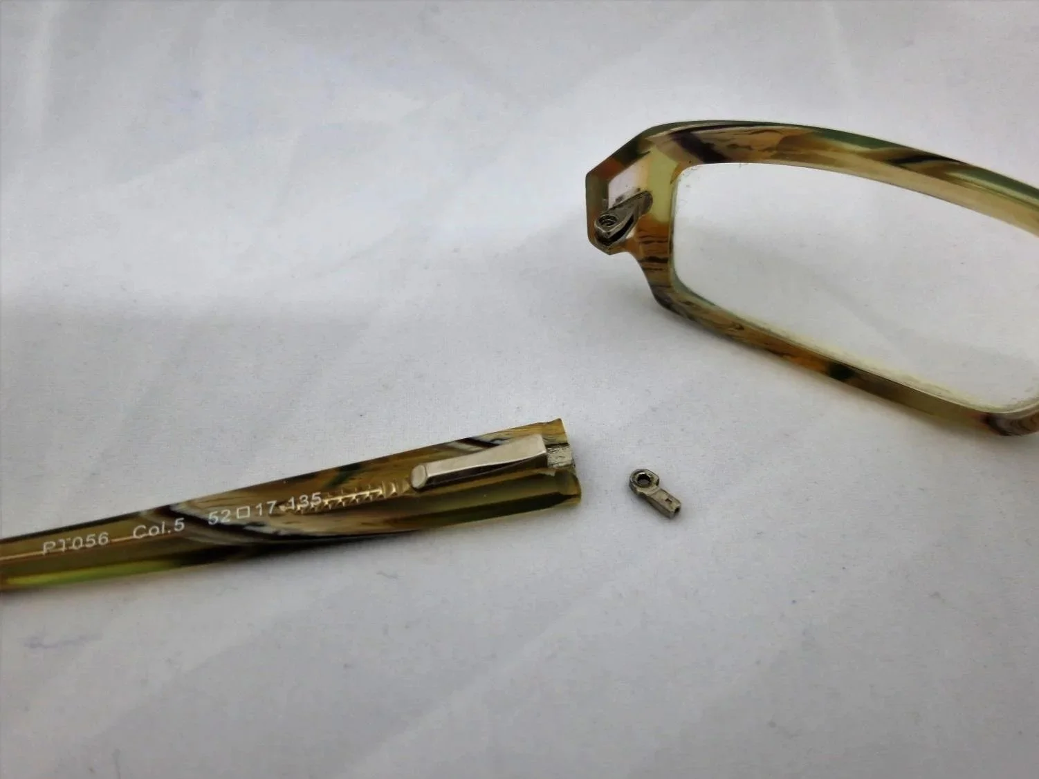 Disassembled pair of eyeglasses with tortoiseshell frame, lens, and small screw on a white surface.