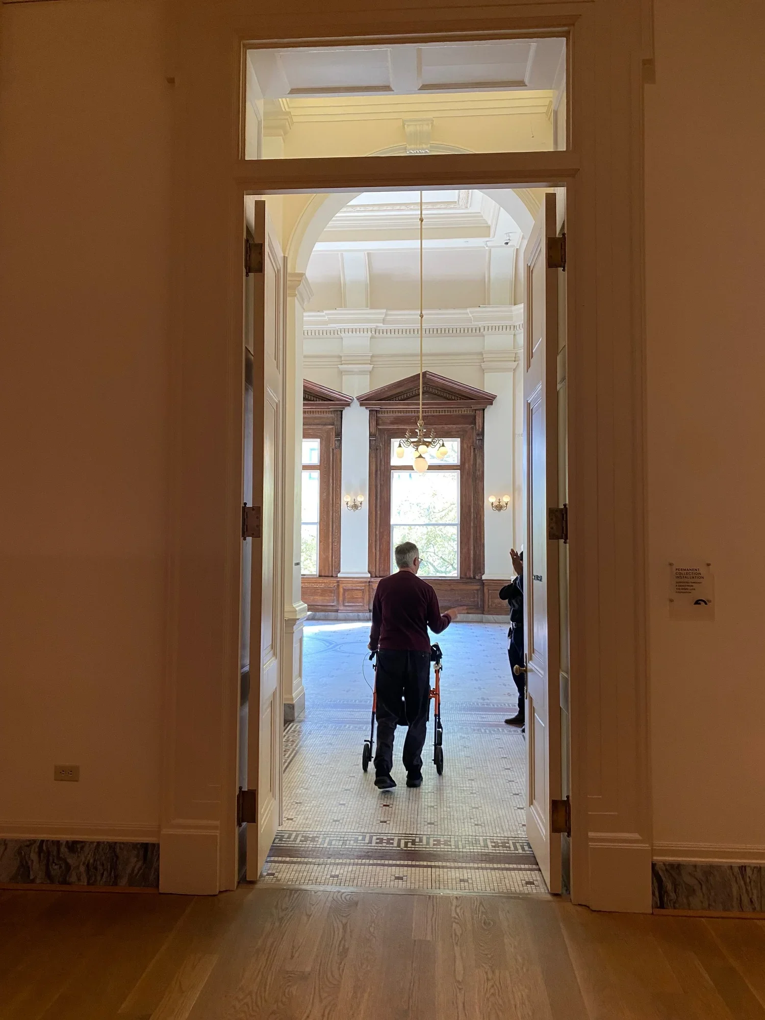 A view through a very tall doorway of a man using a rollator standing in the rotunda on the other side of the open french doors.