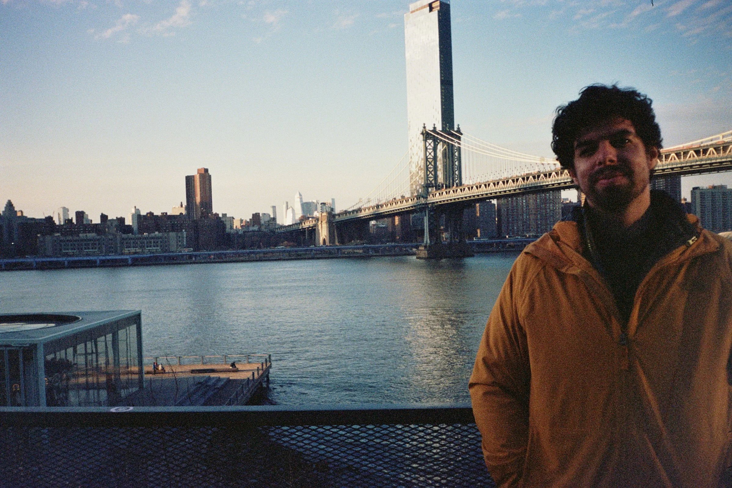 sideOUTsticks CEO, Yves Fogel, standing near waterfront with a city skyline and Brooklyn Bridge in the background, on a clear day.