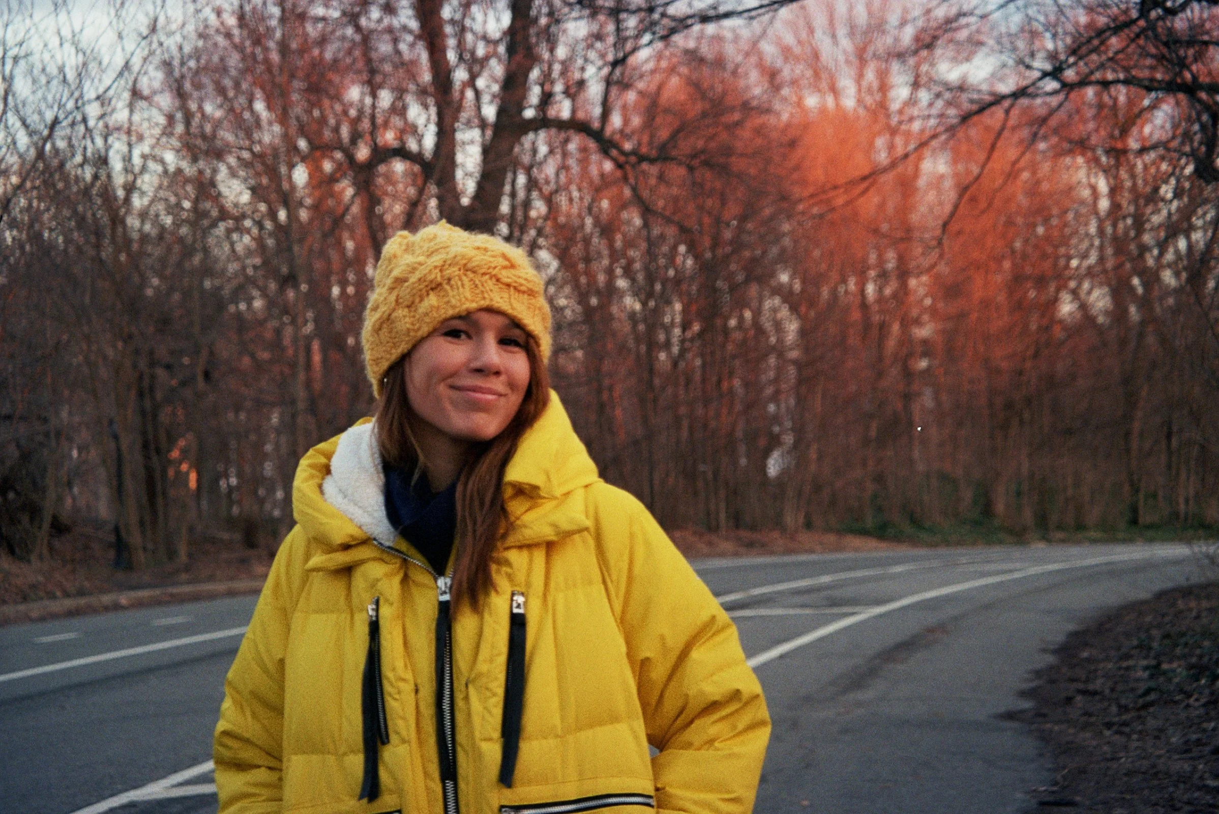 sideOUTsticks Head of Marketing, Romina Larrosa, wearing a yellow jacket and matching yellow knit hat standing on a winding road surrounded by trees with autumn foliage.