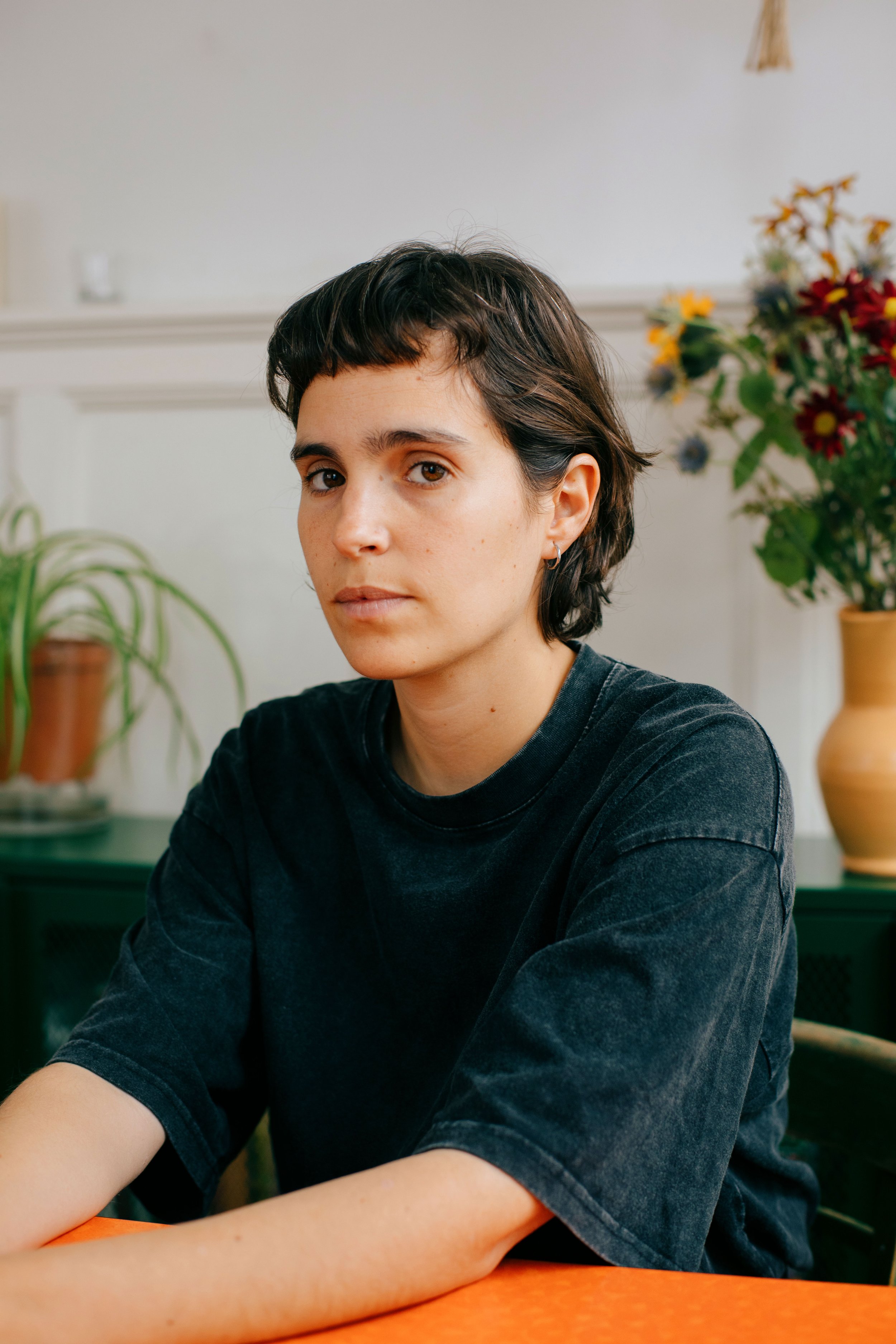 sideOUTsticks AI director, Catalina Torres, wearing a black t-shirt, sitting at a table with a green cabinet and a vase of colorful flowers in the background.