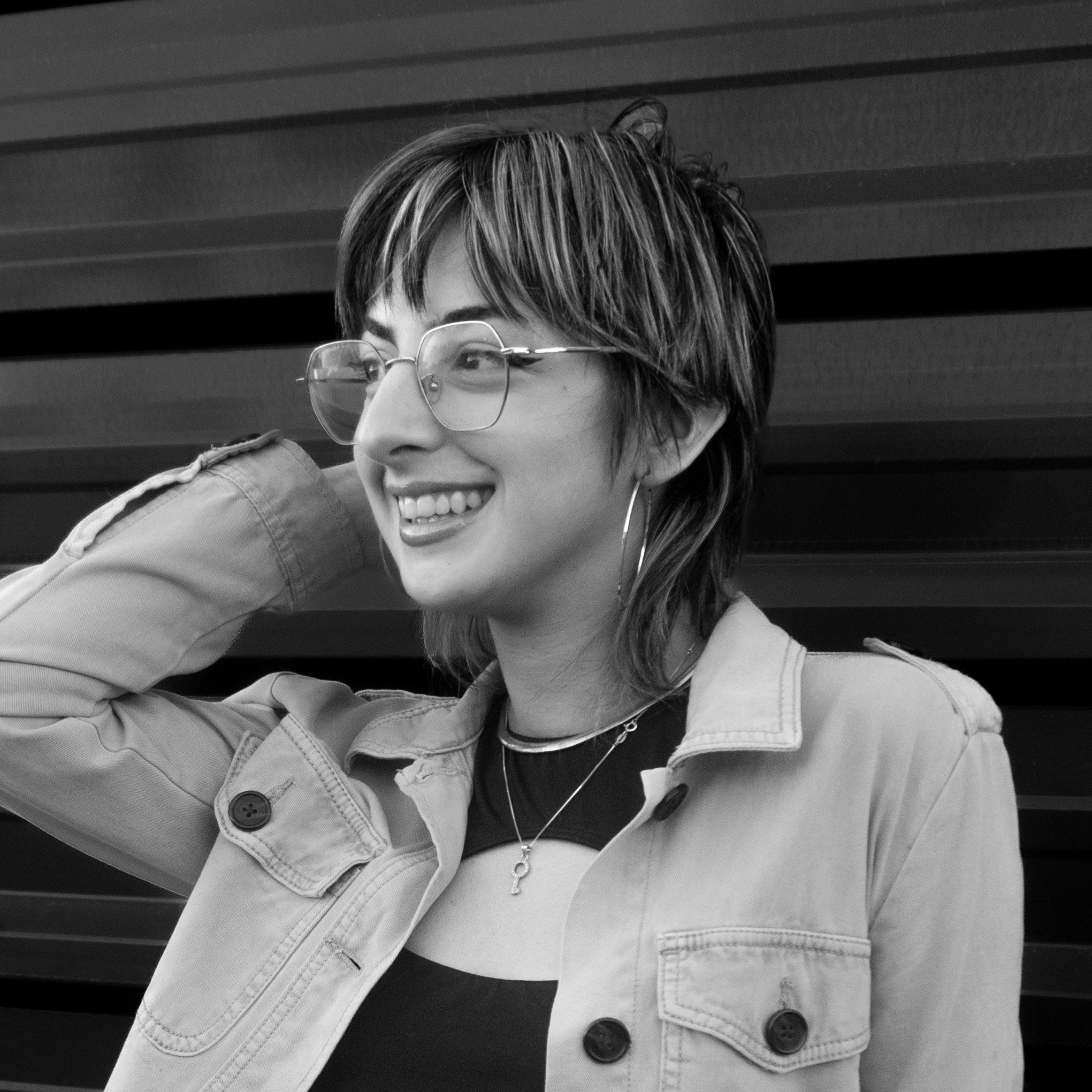 Black and white photo of a young woman with short, layered hair, wearing glasses, earrings, and layered necklaces. She is smiling and looking to her right, with her left hand resting behind her head. She's wearing a light-colored jacket over a dark top, with a patterned background.