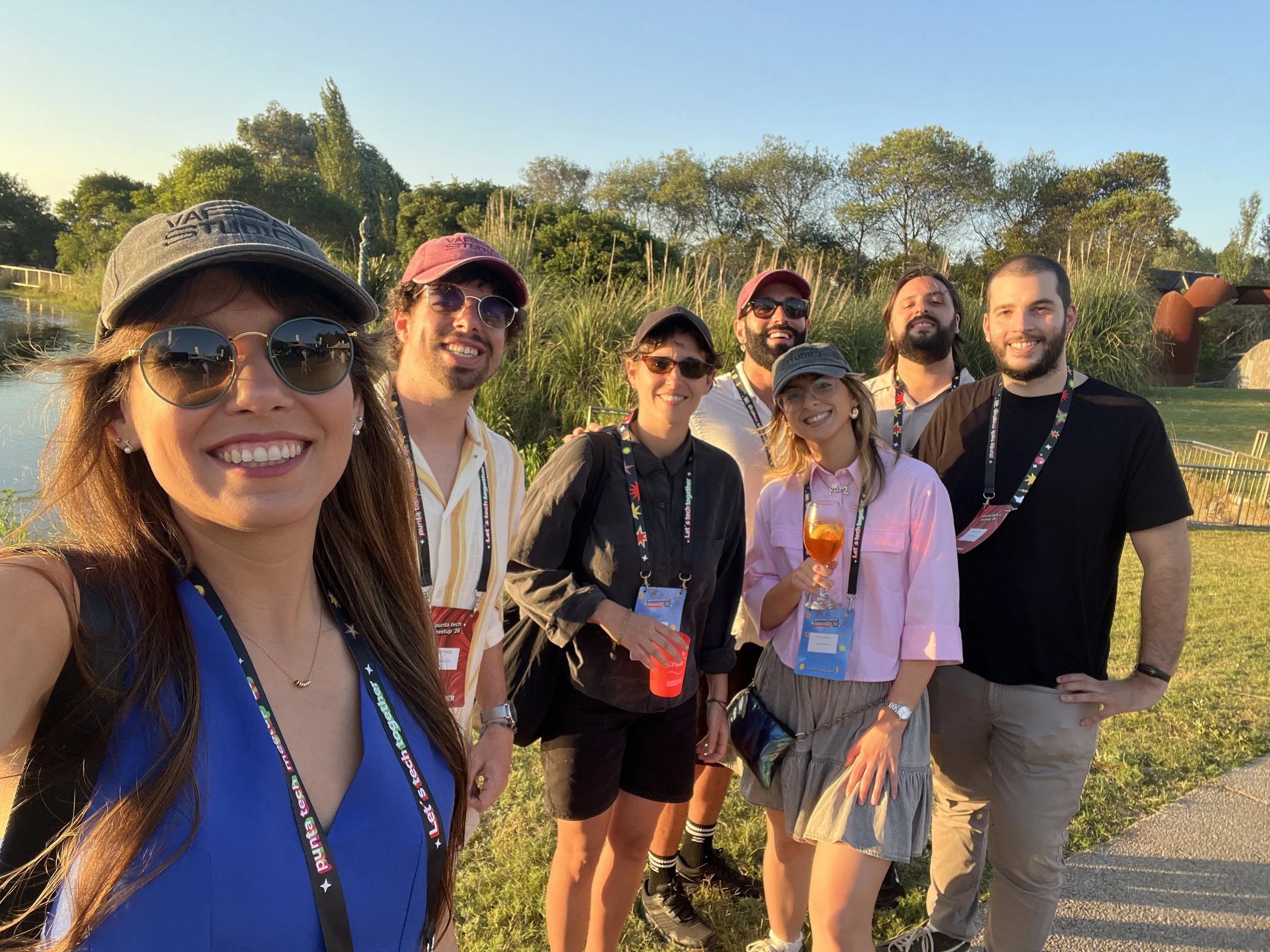 The sideOUTsticks team. A group of eight diverse people smiling outdoors during sunset, wearing casual clothing, some with sunglasses and lanyards, standing on grass near a water feature with trees and modern sculptures in the background.