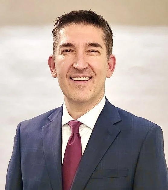 A man smiling in a navy blue suit, white shirt, and maroon tie, with short dark hair, standing indoors against a blurred background.