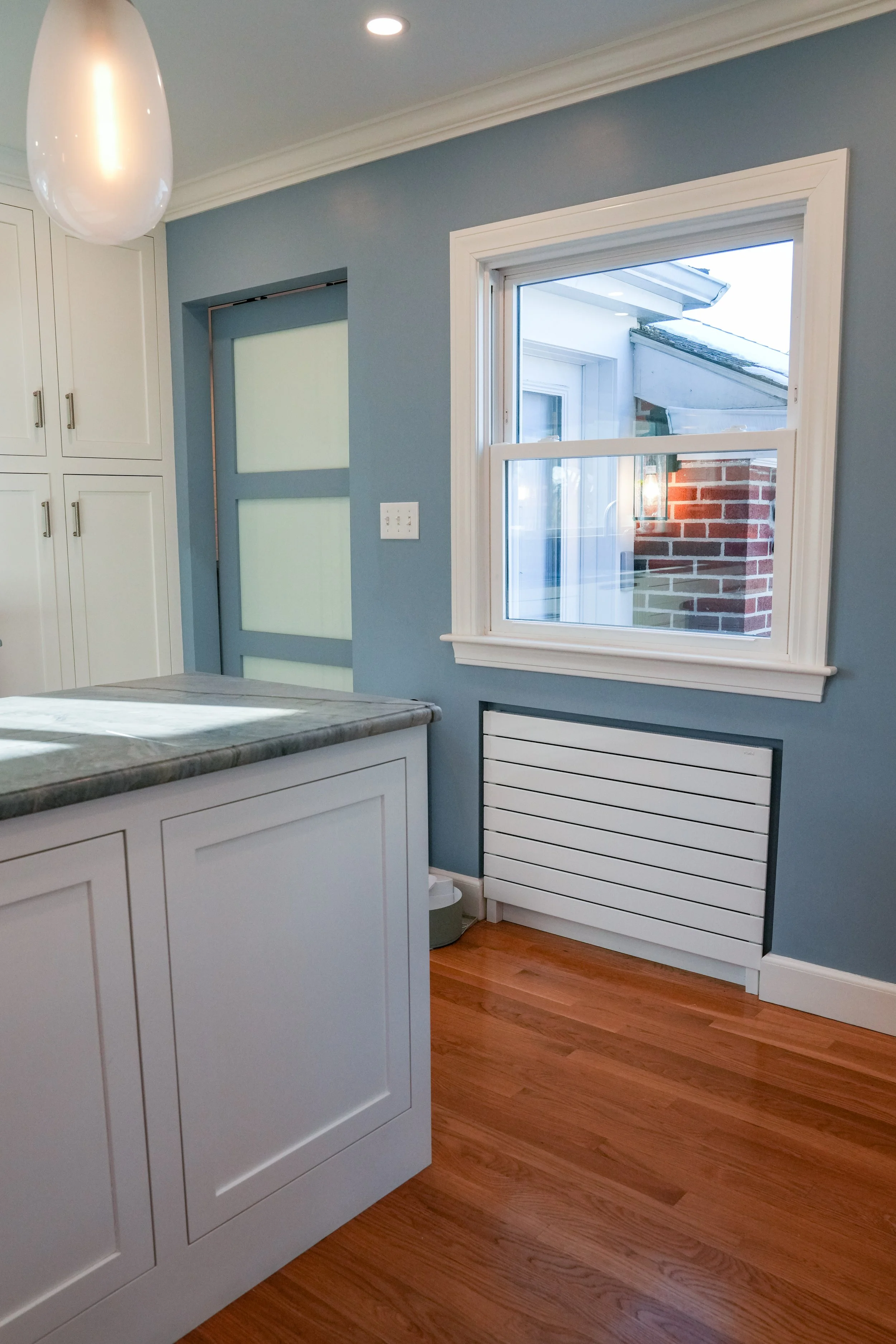Kitchen with white cabinets, a marble countertop, a blue wall, a window showing a brick wall outside, and a white radiator below the window.