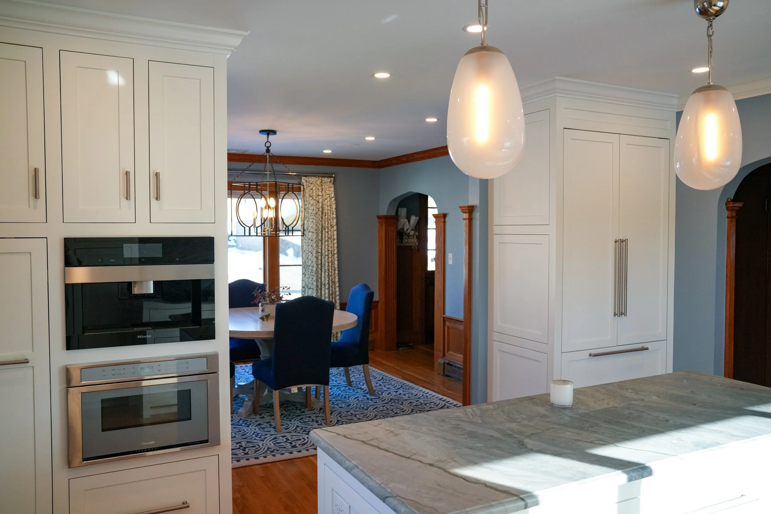 A kitchen and dining area with white cabinetry, a marble island, pendant lighting, and a dining table with blue chairs. There's a window with curtains and a chandelier in the background.