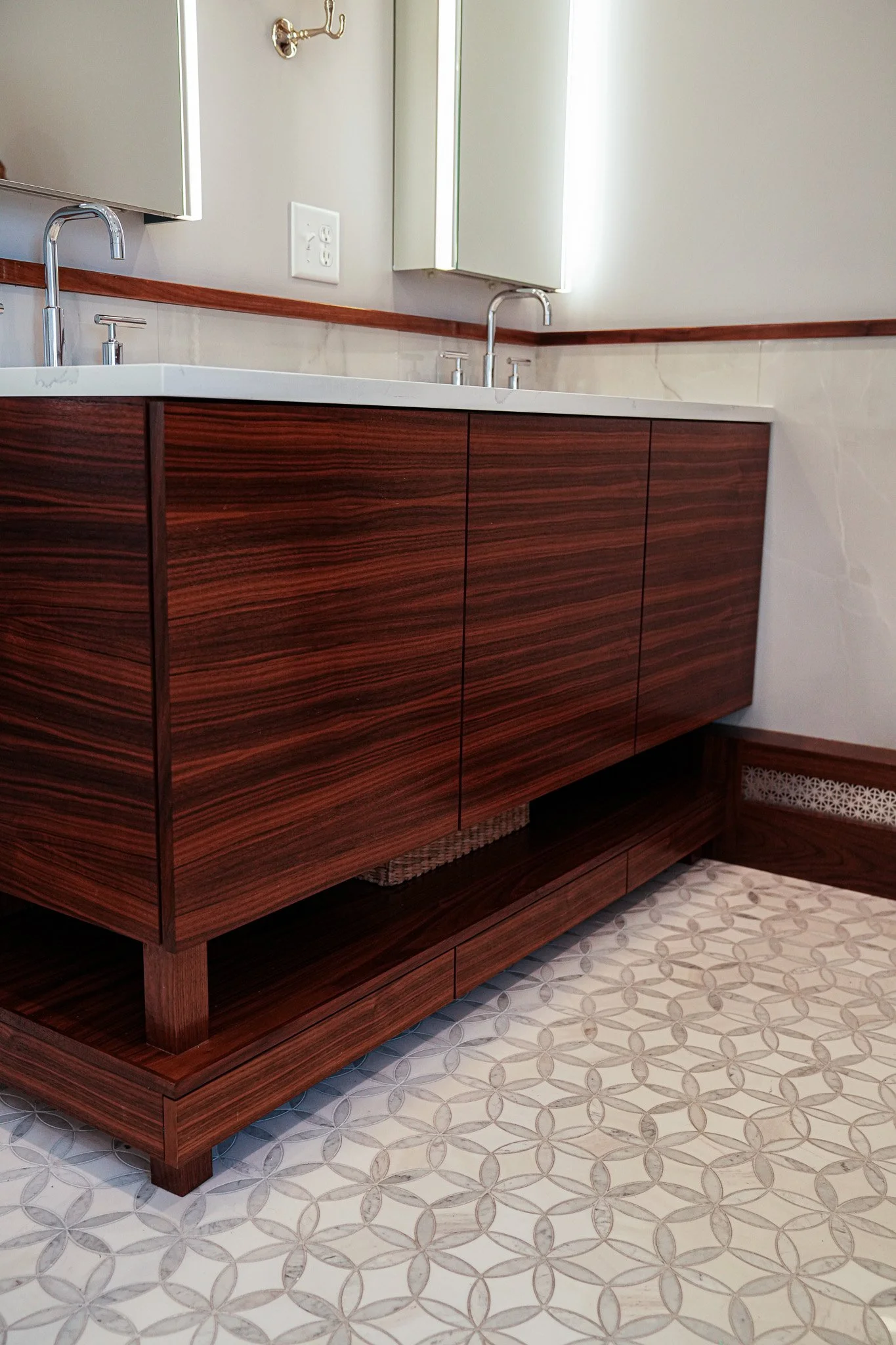 Wooden bathroom vanity with a white countertop and undermount sink, with two faucets, set against a tiled wall with a small mirror and a wall hook, and a geometric patterned floor.
