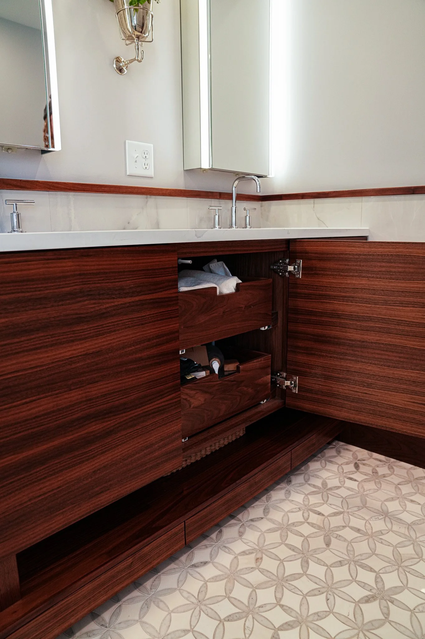 A bathroom vanity with a wood finish, a white countertop, and a faucet. The vanity has an open cabinet with towels and other items inside. The wall behind has a mirror, a small cabinet, and a light fixture. The floor has a patterned tile design.