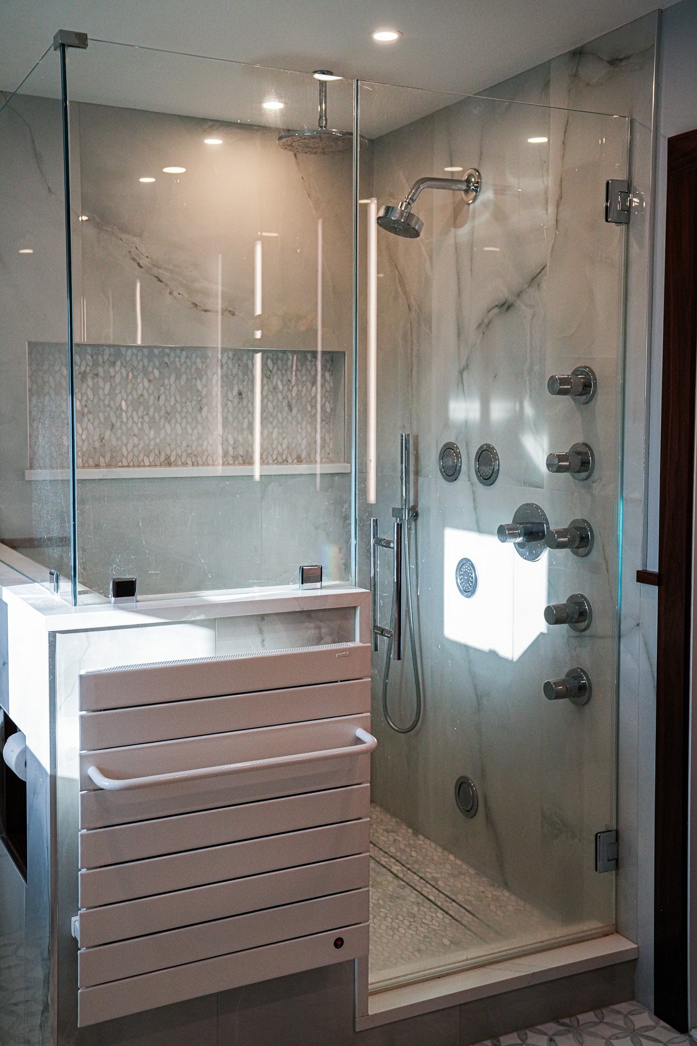 A modern shower with a glass enclosure, marble walls with subtle veining, a rainfall showerhead, multiple body jets, a handheld shower, and a textured tile floor.