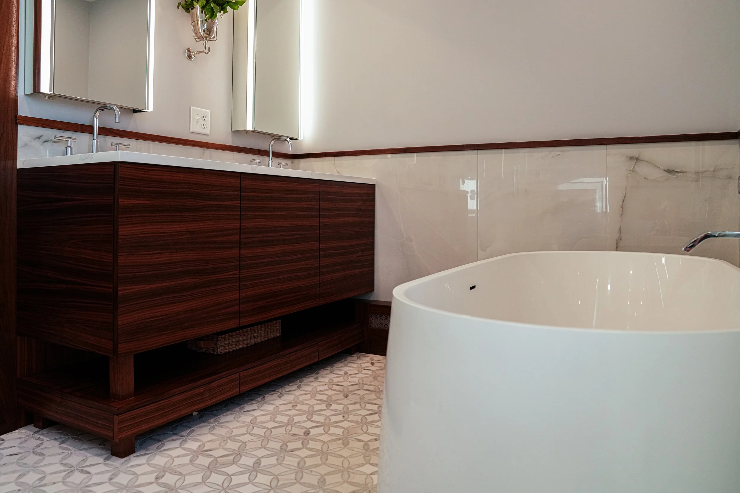 A bathroom with a wooden vanity, two sinks, a mirror, and a bathtub. The wall is partially tiled with marble, and the floor has patterned tiles. There is a plant hanging above one of the sinks and four electrical outlets on the wall.