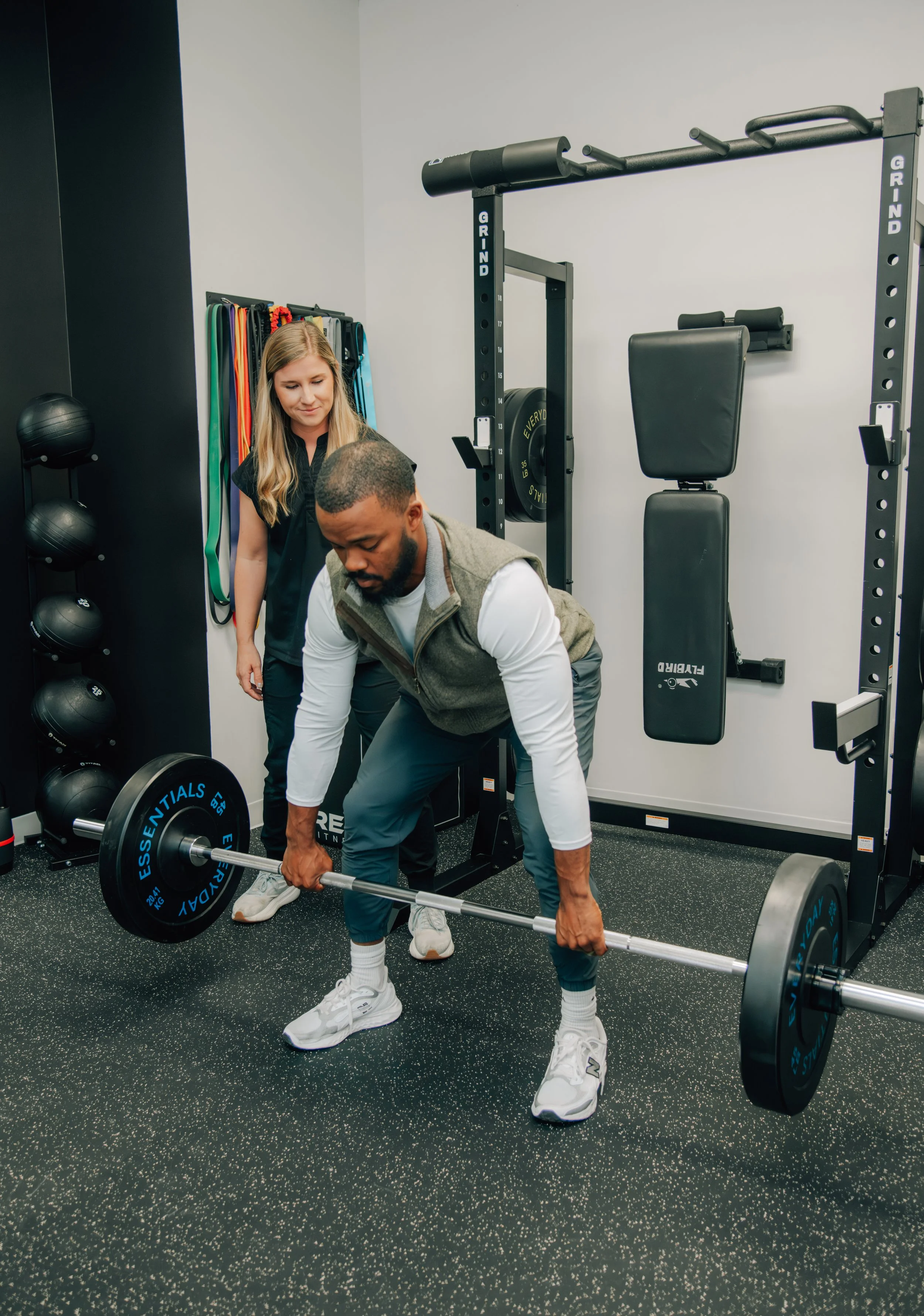 A man lifts a barbell while a woman supervises at a gym.