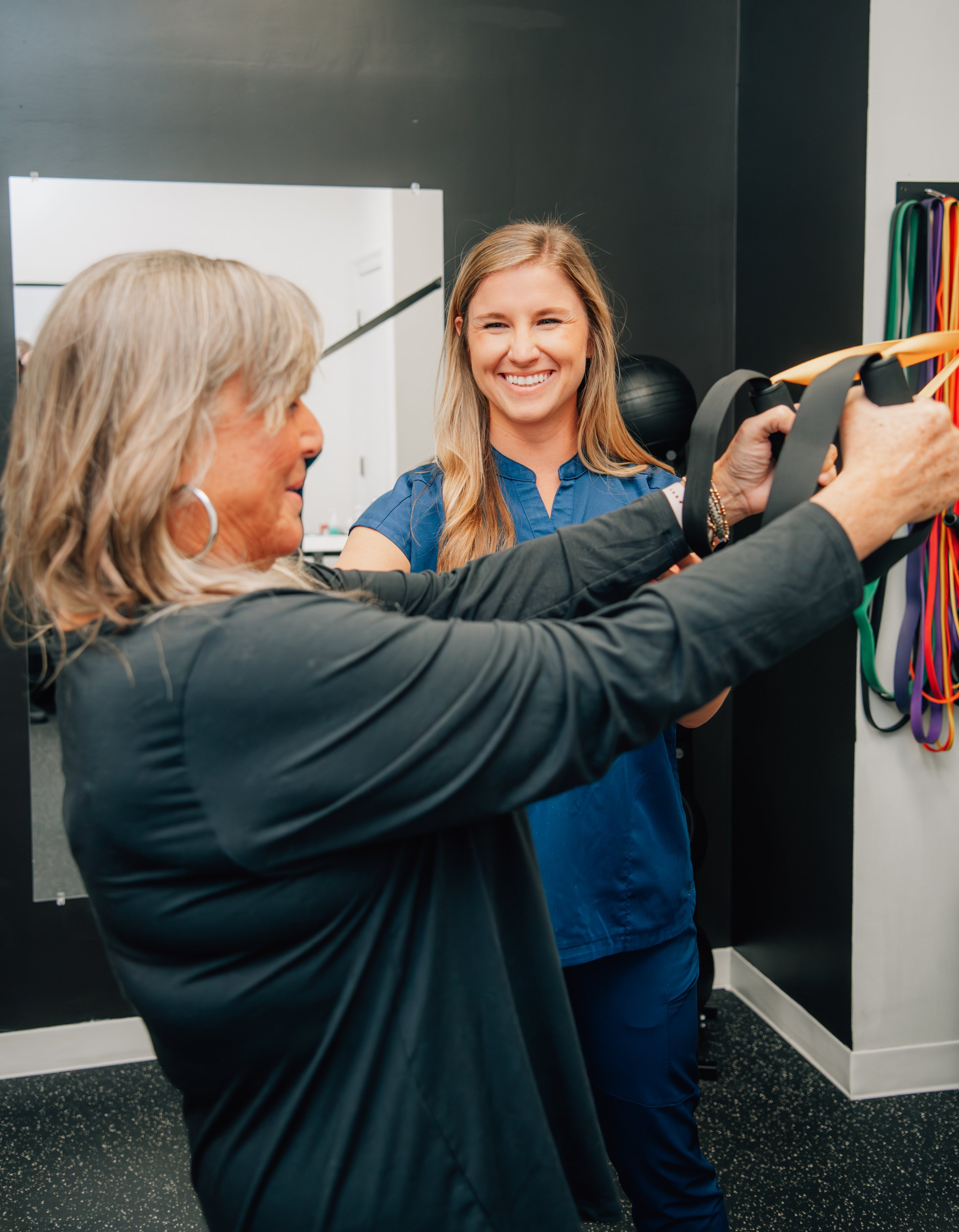 A young woman in blue scrubs smiling as an older woman demonstrates exercises with resistance bands in a gym or fitness studio.