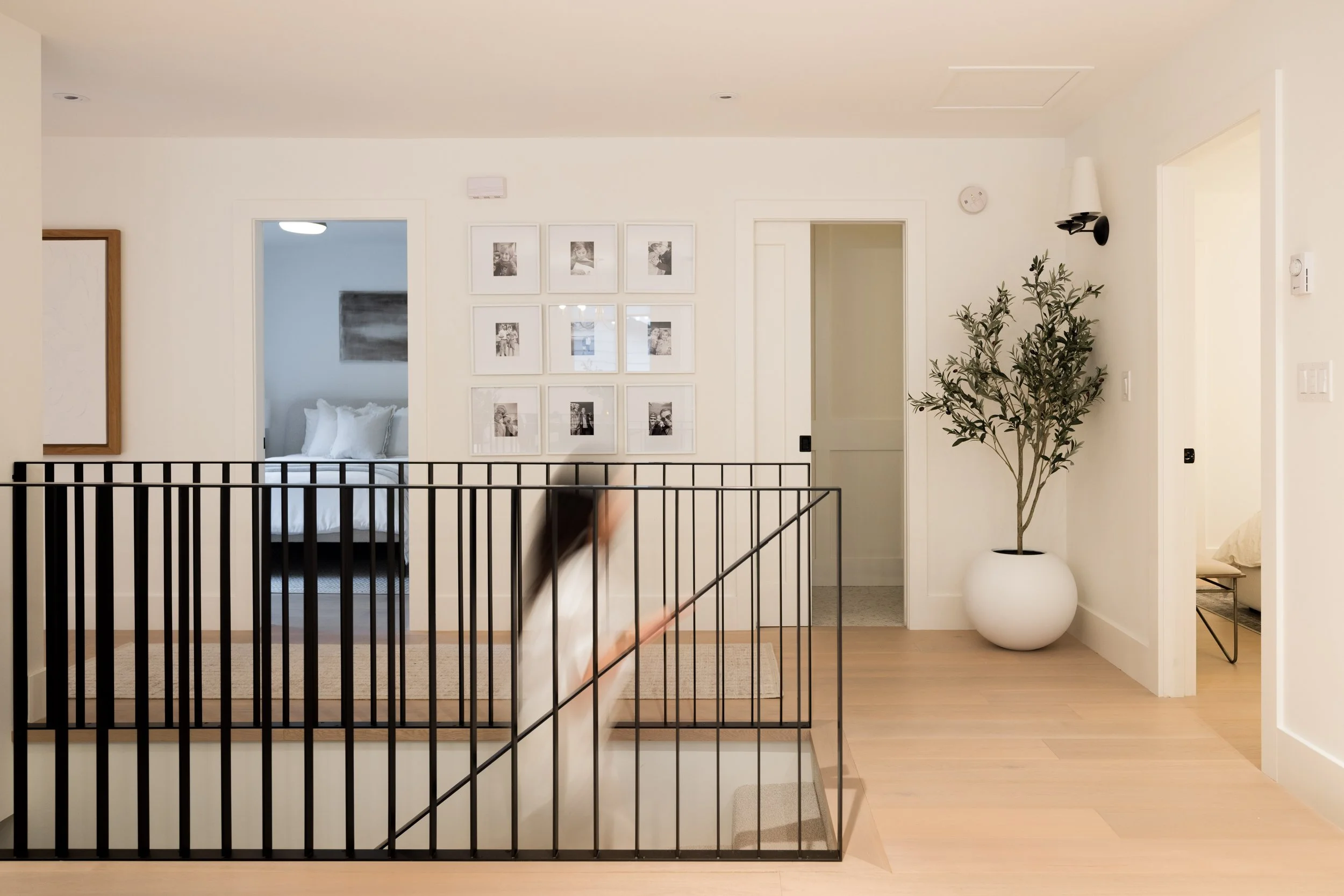 Interior of a modern home with a staircase, framed photos on the wall, a large potted plant, and two bedrooms visible through doorways.