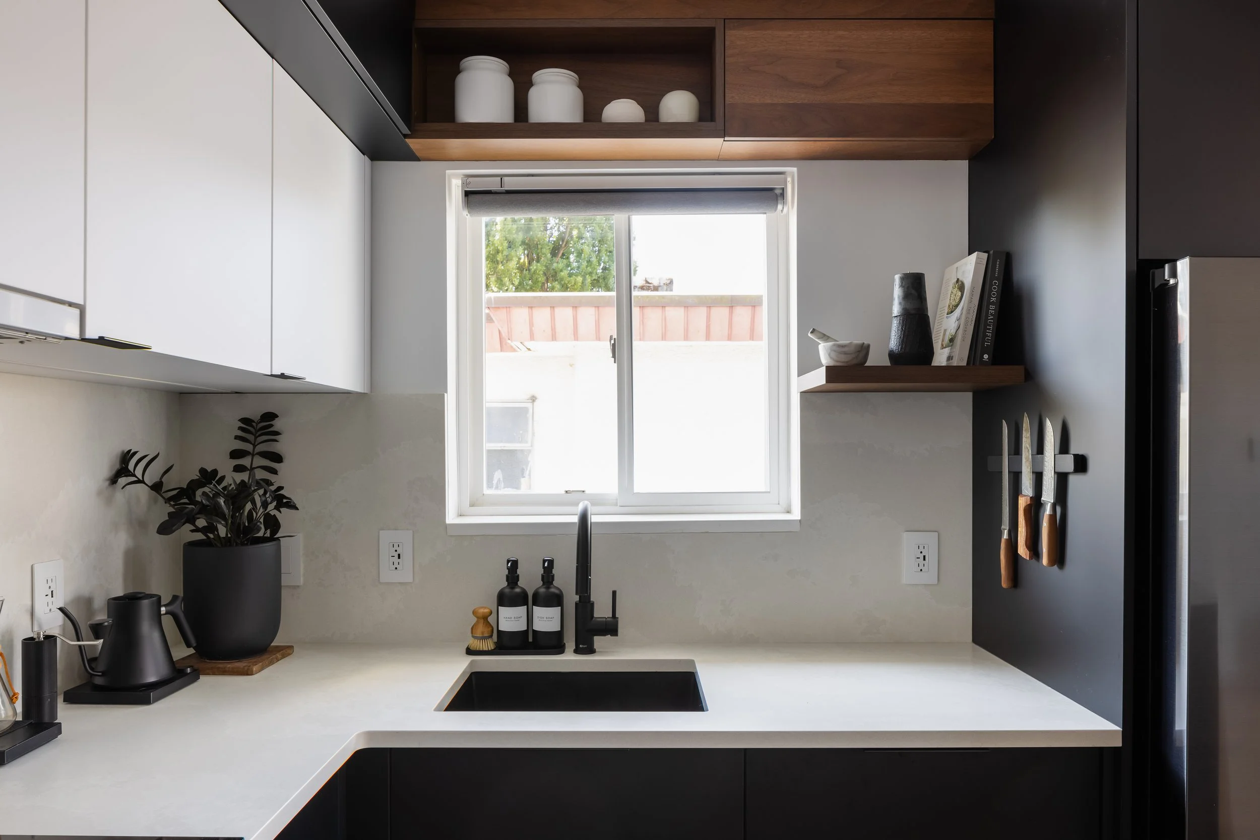 Modern kitchen with black and white cabinetry, a white countertop, a black sink, and a window with a view outside. Open shelves with white jars and decorative items, and knives hanging on the wall.