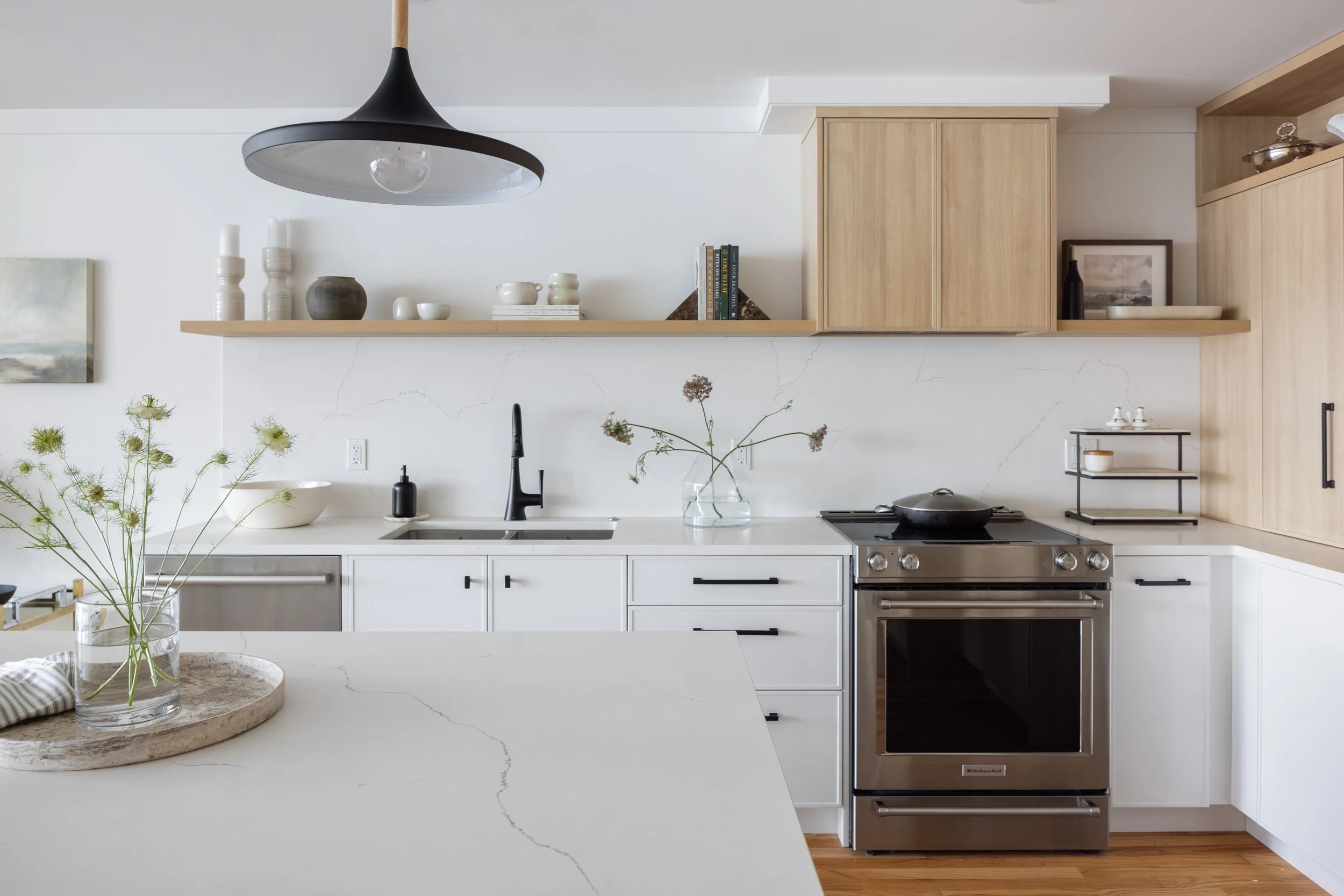 Modern kitchen with white countertops, stainless steel oven, and light wood cabinets. Open shelf with decorative items and a plant on the counter.