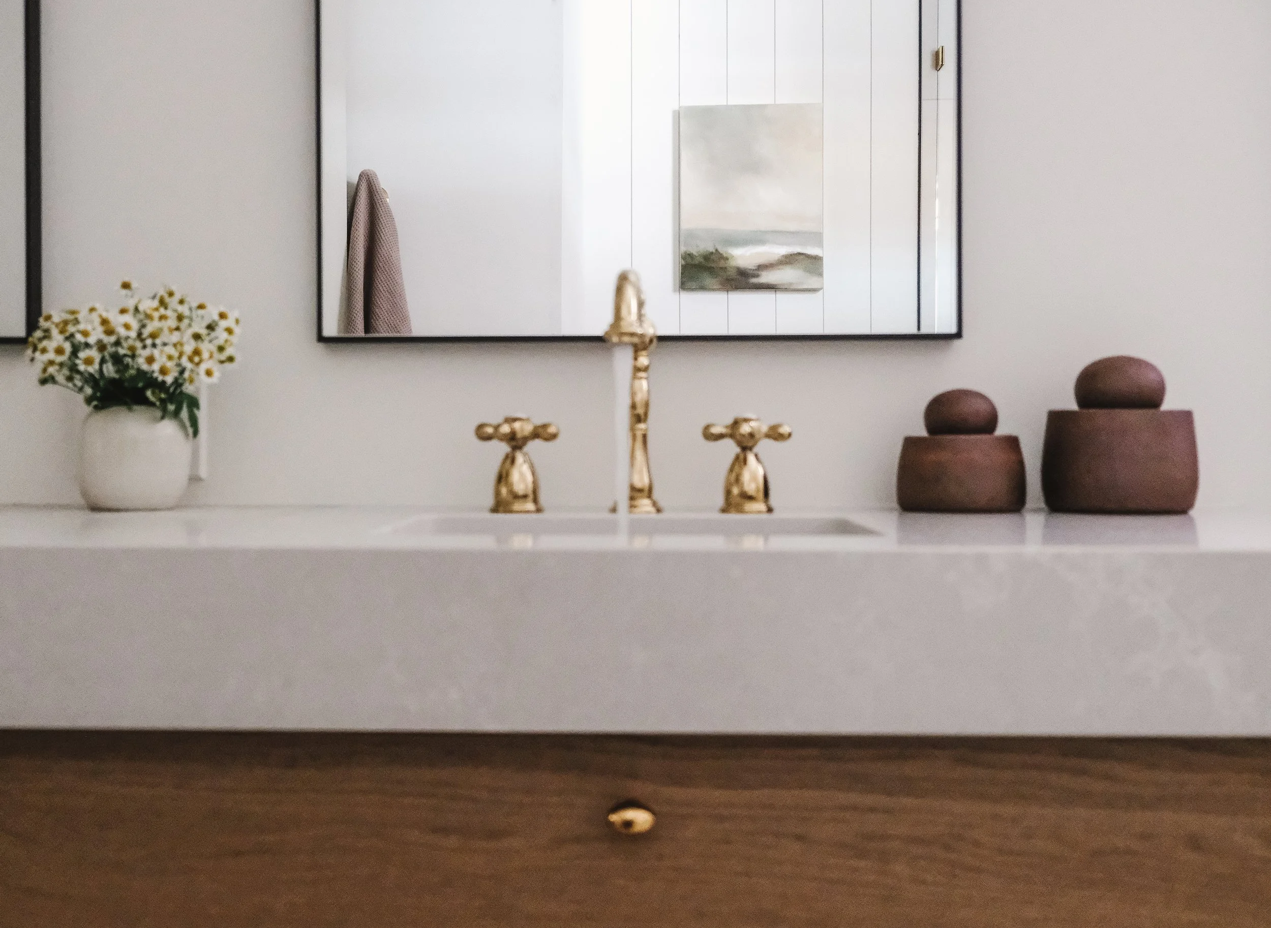 Bathroom vanity with a vase of white daisies, a mirror, a painting, gold faucet & handles, and decorative wooden boxes.