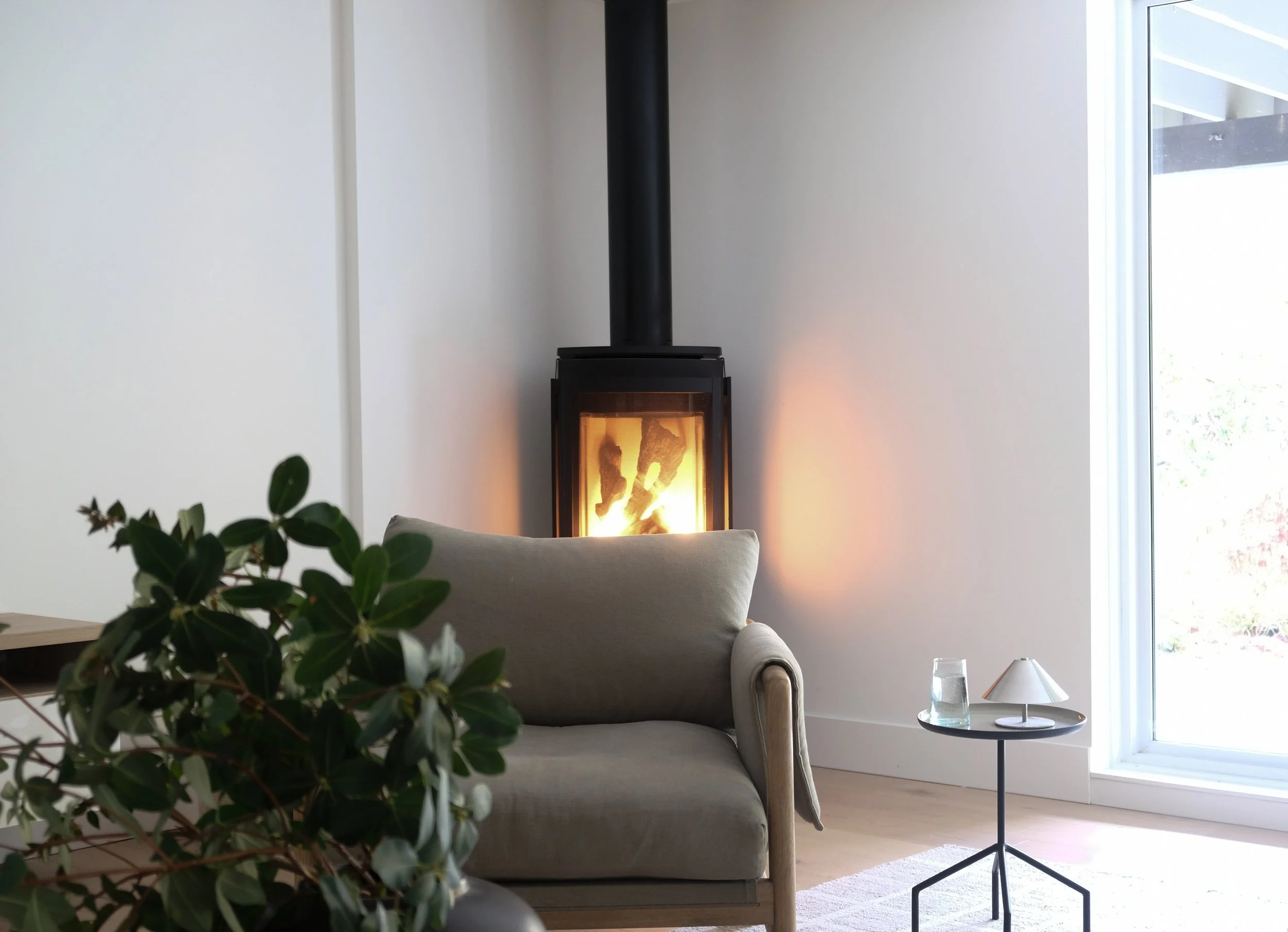 Living room with a beige sofa, a small black side table with a glass of water and a lamp, a white wall, a window showing outdoors, and a modern black wood-burning stove with a visible fire inside.