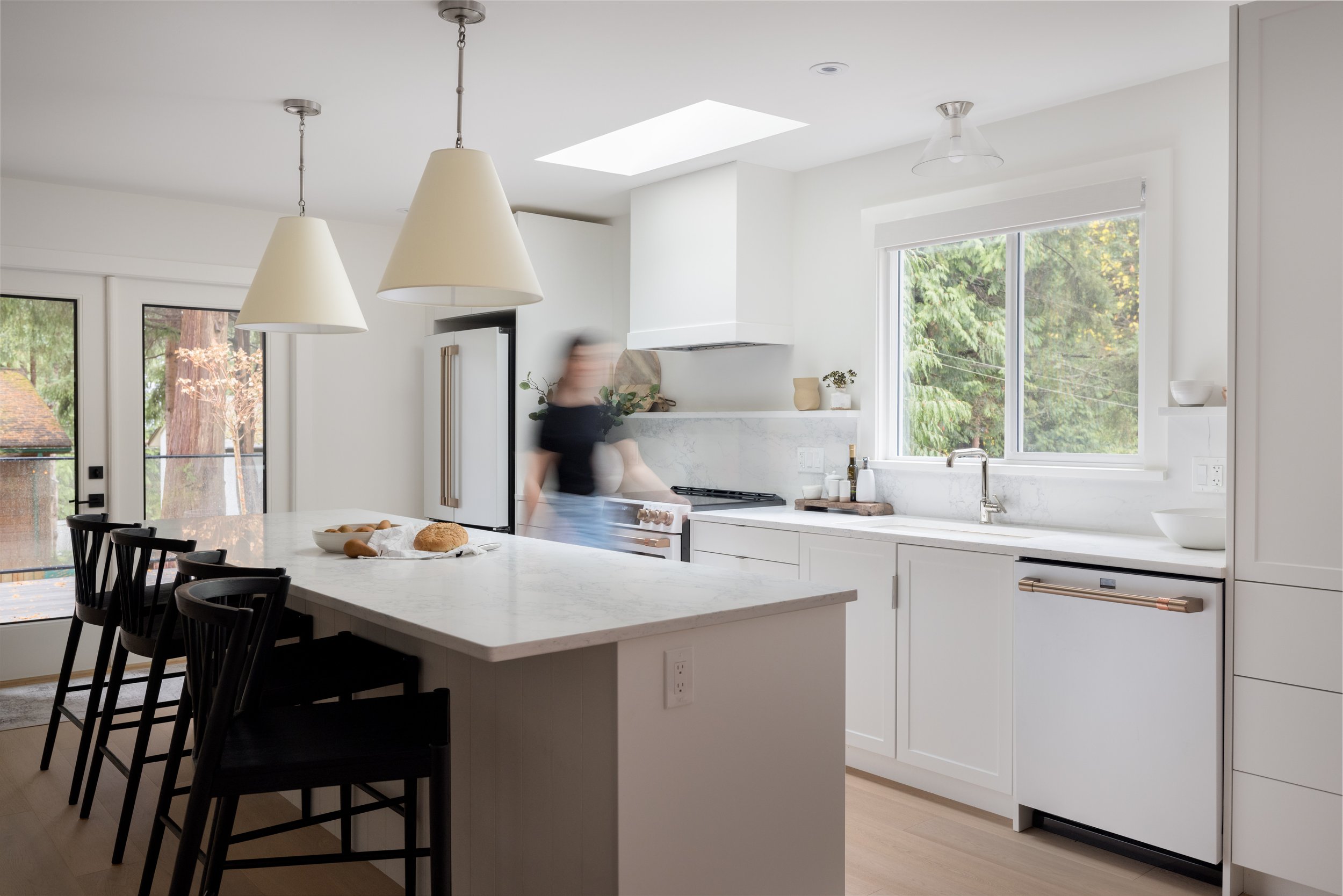 Modern white kitchen with island, black bar stools, large window showing greenery, and a person moving near the stove
