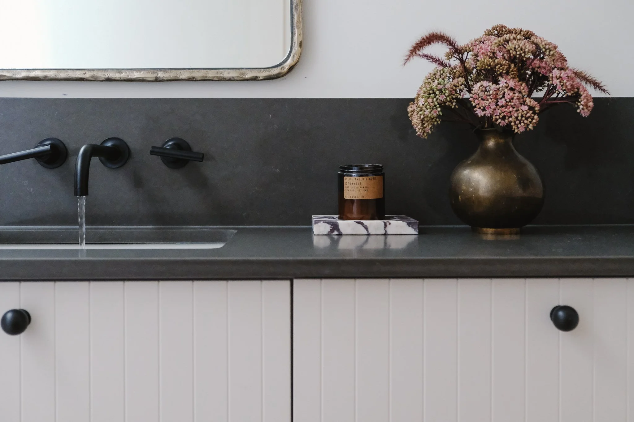 A bathroom countertop with a black faucet, a brown candle labeled 'Amber & Moss,' a marble tray, and a bronze vase with pink and purple flowers.