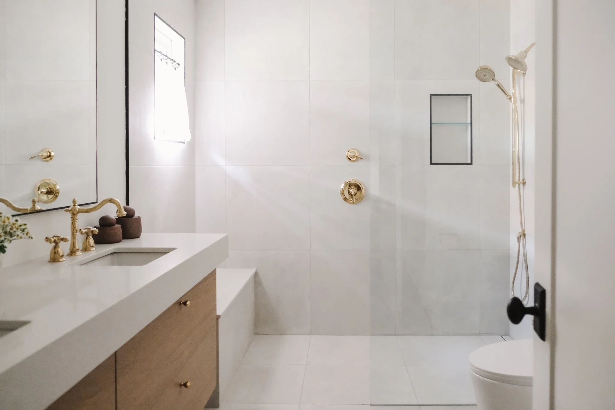 Modern bathroom with white walls and floor, gold fixtures, a window, a vanity with a sink, mirror, and decor, and a walk-in shower with a handheld showerhead and built-in niche.