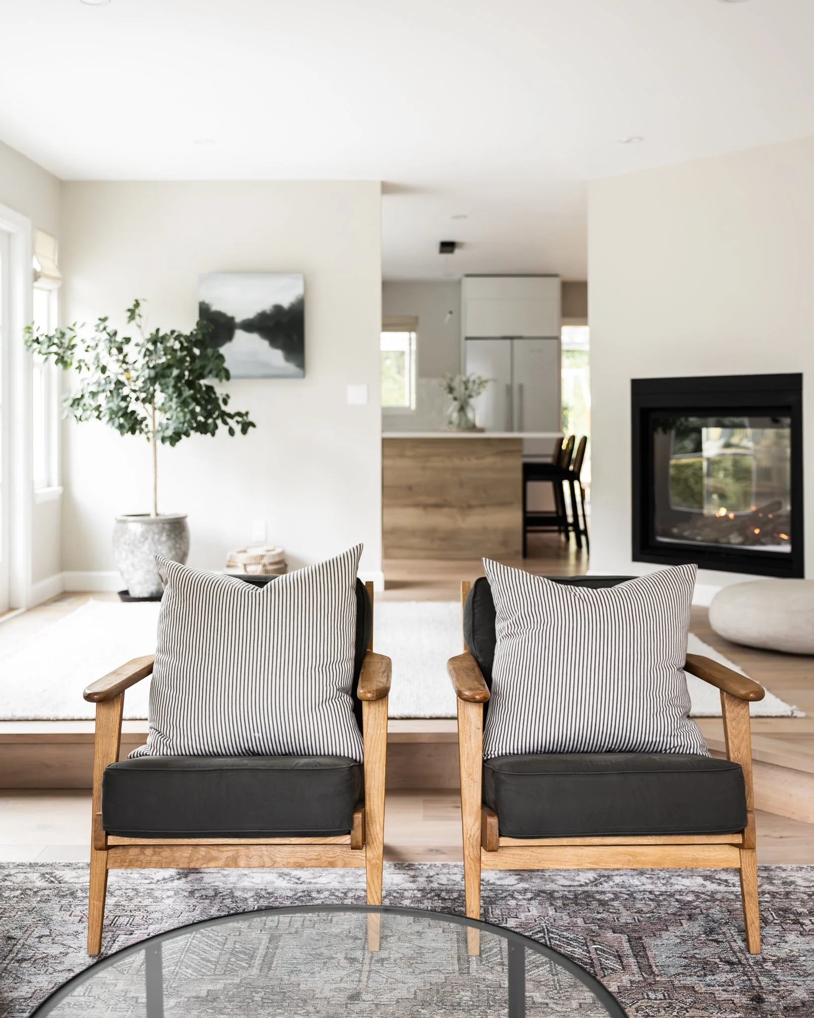 Living room with two wooden armchairs with black cushions and striped pillows, a modern fireplace, a potted plant, and a white rug on wooden flooring.