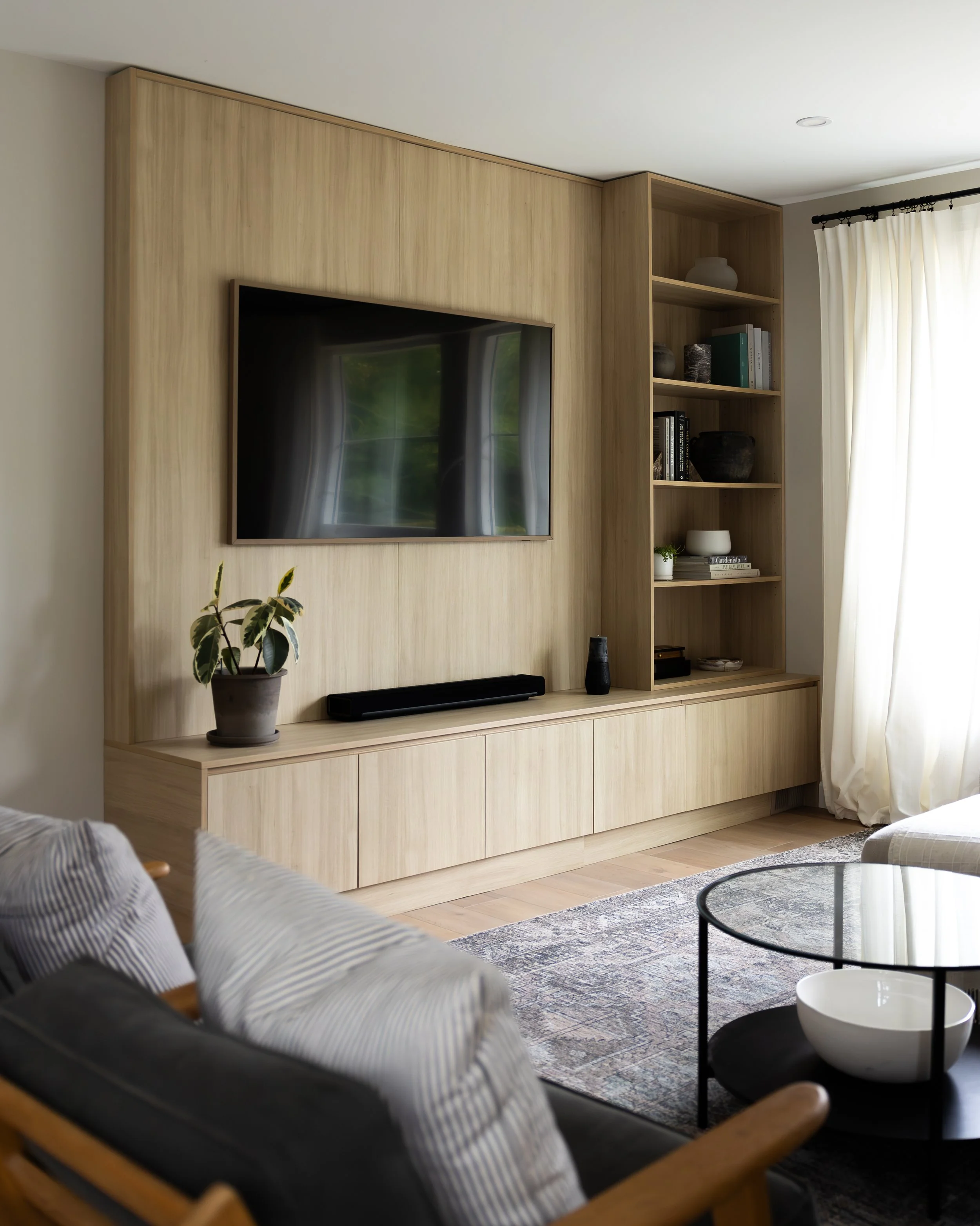 Modern living room with wooden panel wall, mounted flat-screen TV, open shelving with books and decor, potted plant, and a large window with cream curtains.
