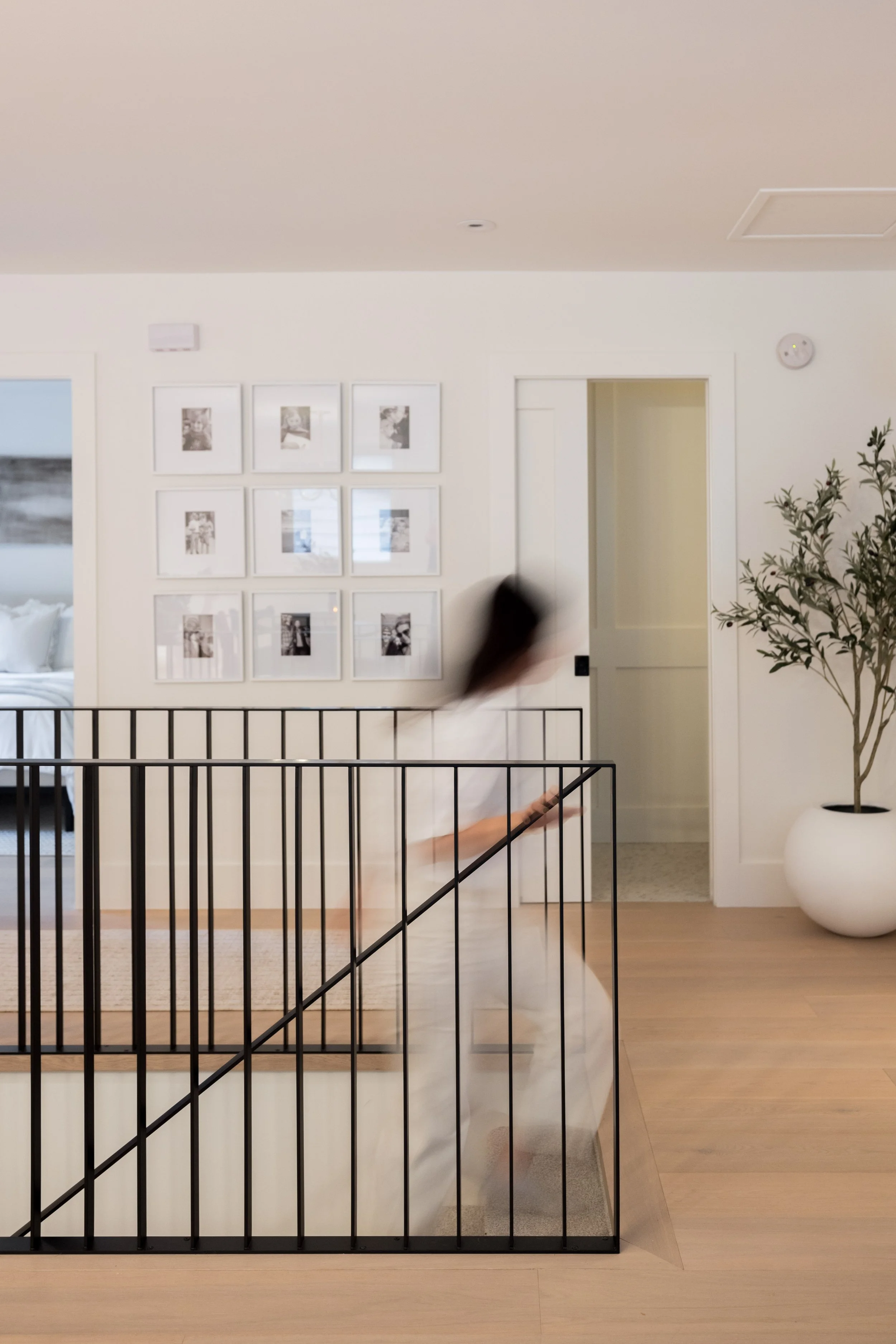 A person descending a staircase inside a modern home with a black railing, white walls, framed pictures, a potted tree, and a doorway.