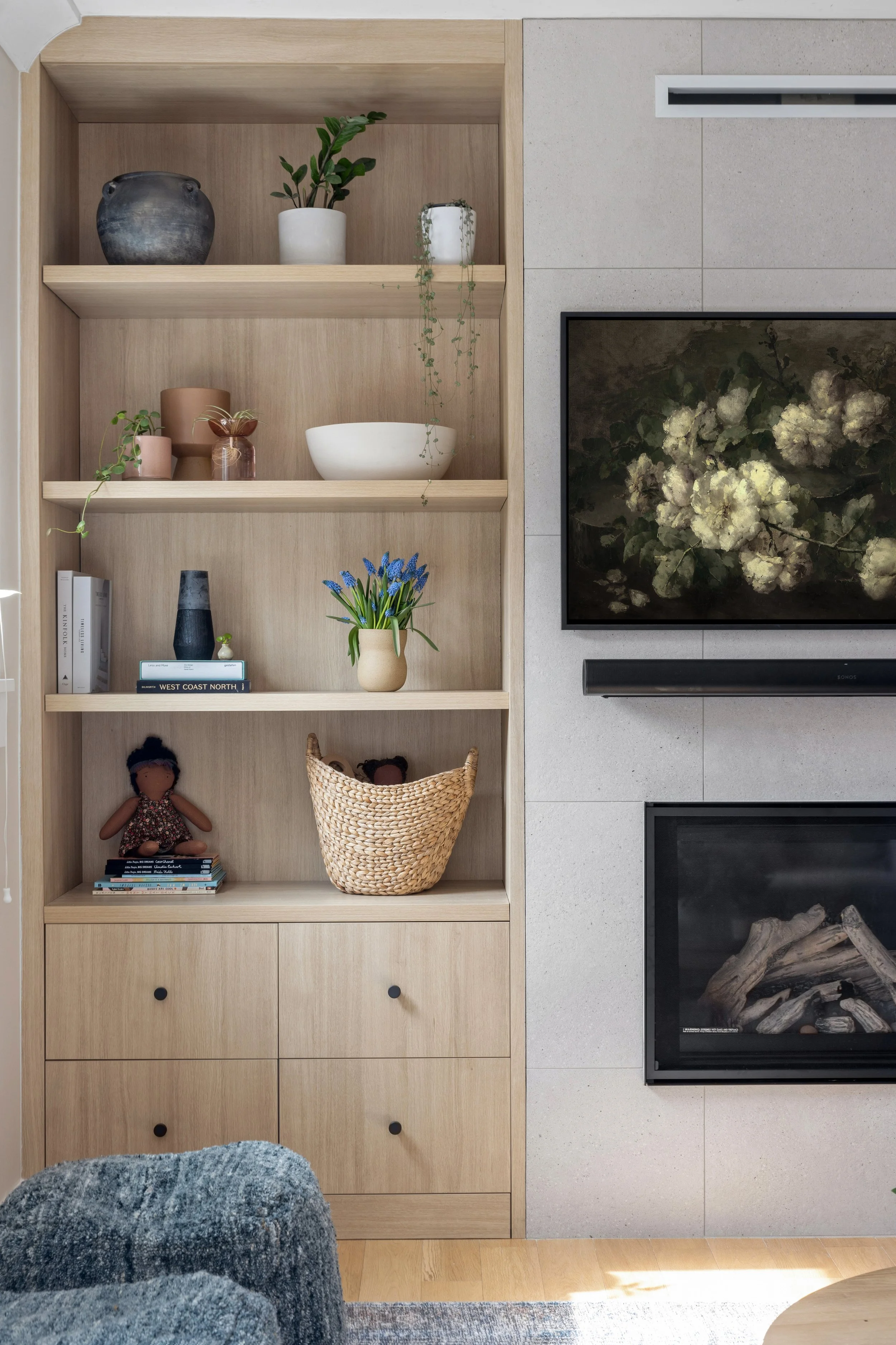 A built-in wooden bookshelf filled with decorative objects, books, and dolls, situated next to a modern fireplace and television in a living room.
