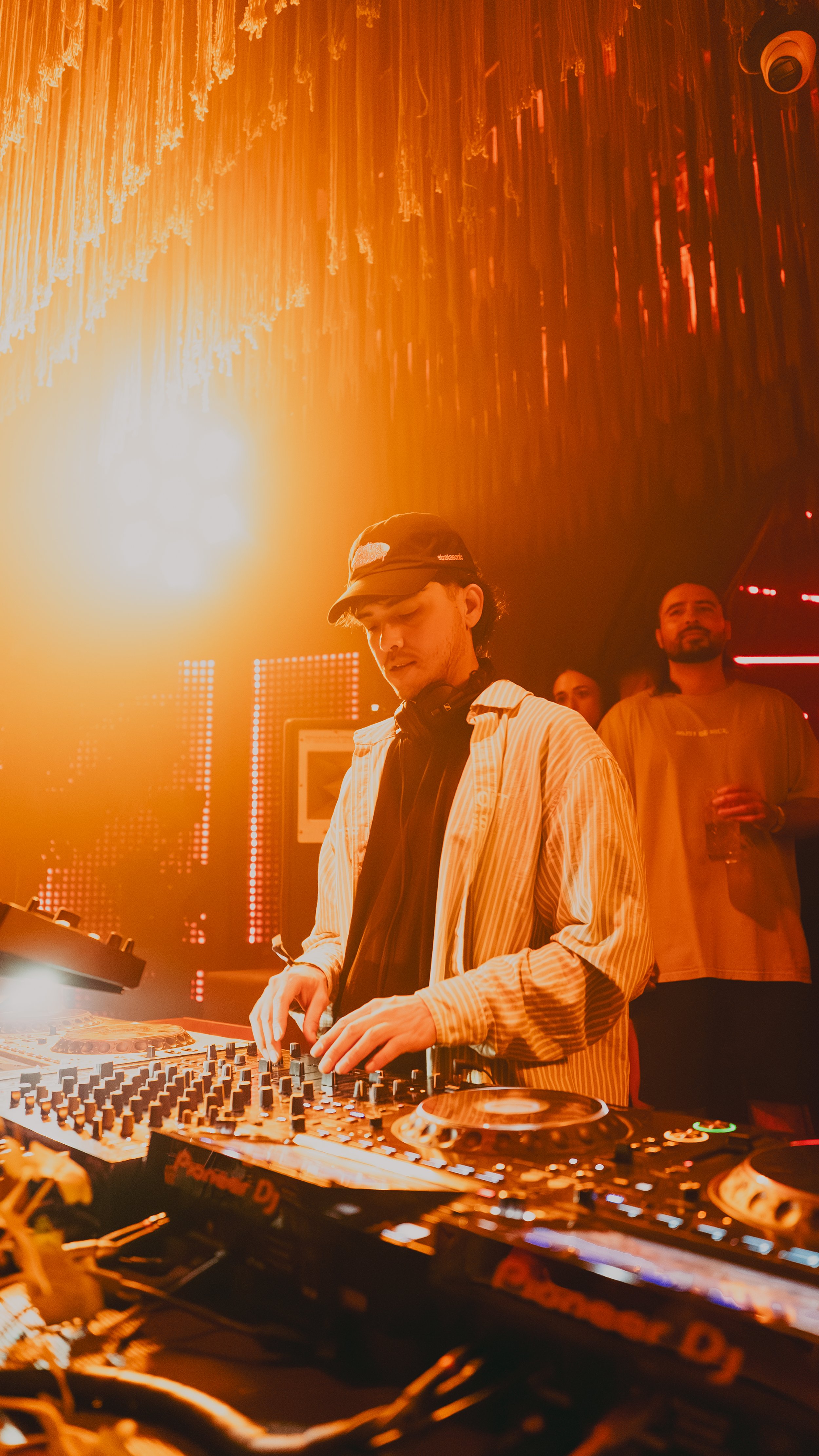 A DJ wearing a striped shirt and a cap is mixing music on a DJ console at a club, with orange lighting and a few people in the background watching and holding drinks.