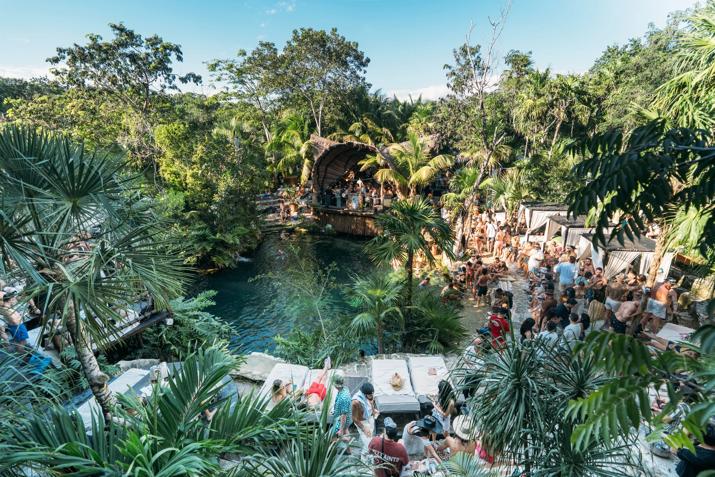 A crowded outdoor pool party surrounded by lush tropical greenery and palm trees, with people relaxing on lounge chairs, swimming, and socializing near a swimming pond and a thatched roof bar.