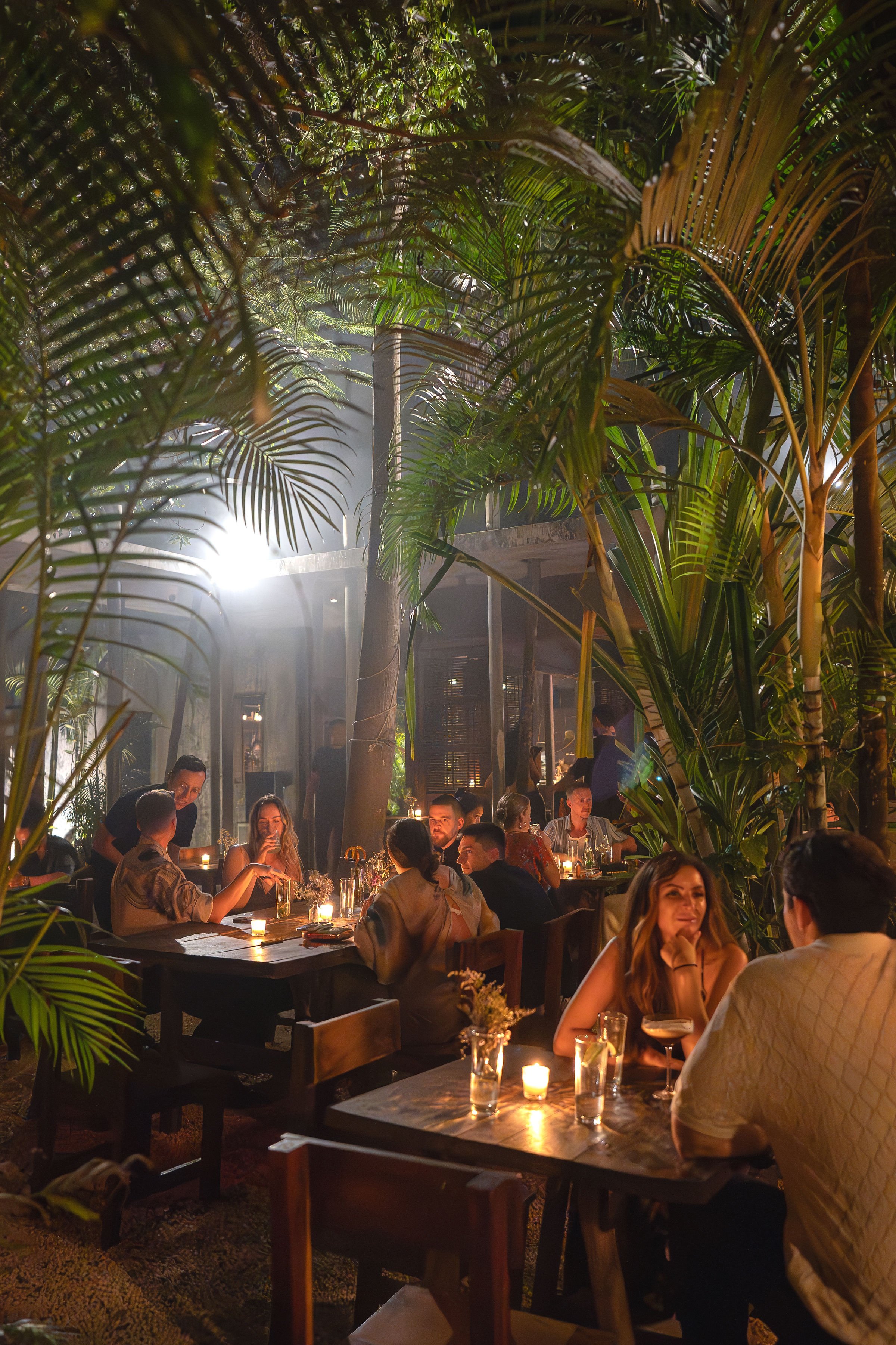 People dining in a tropical-themed restaurant with lush green plants, warm lighting, and candles on tables.