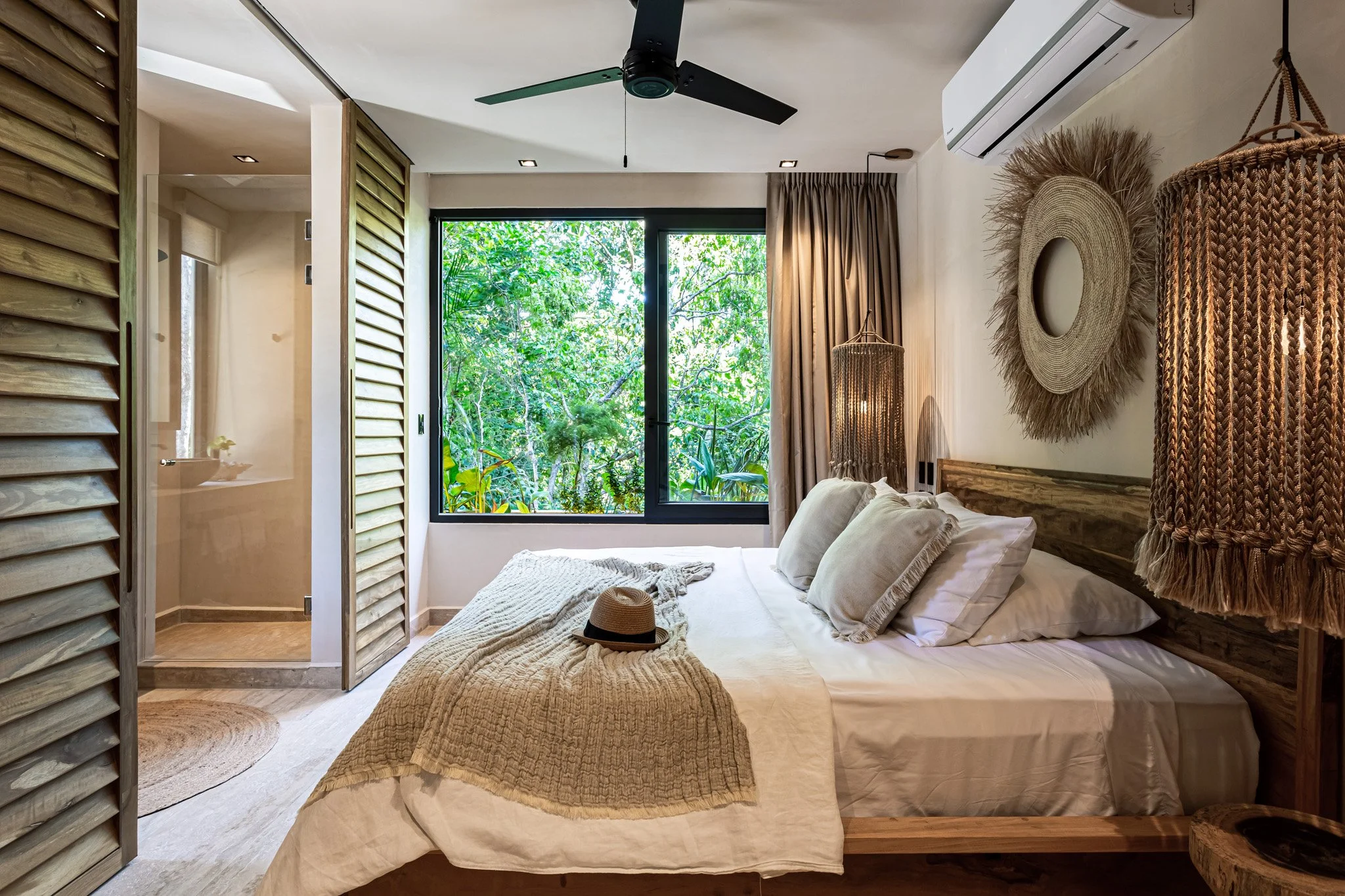 A cozy bedroom with a large bed featuring beige pillows and a beige textured blanket, a sun hat resting on the blanket, a window with green foliage outside, beige curtains, wall decorations including a large woven mirror and basket pendant lights, wo