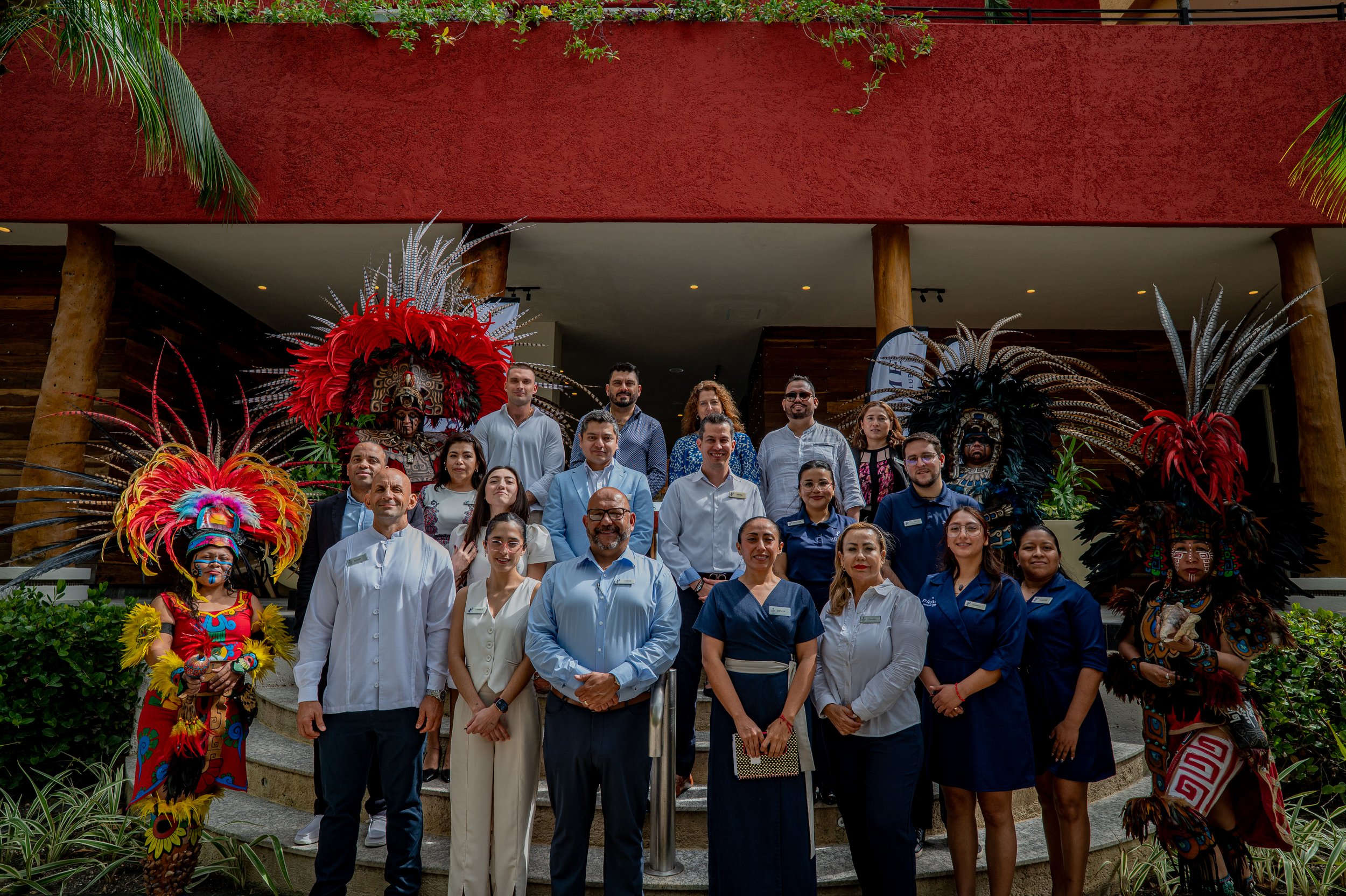 Group of people posing on stairs with traditional dancers in colorful costumes and feathered headdresses, inside a building with wooden pillars and plants.