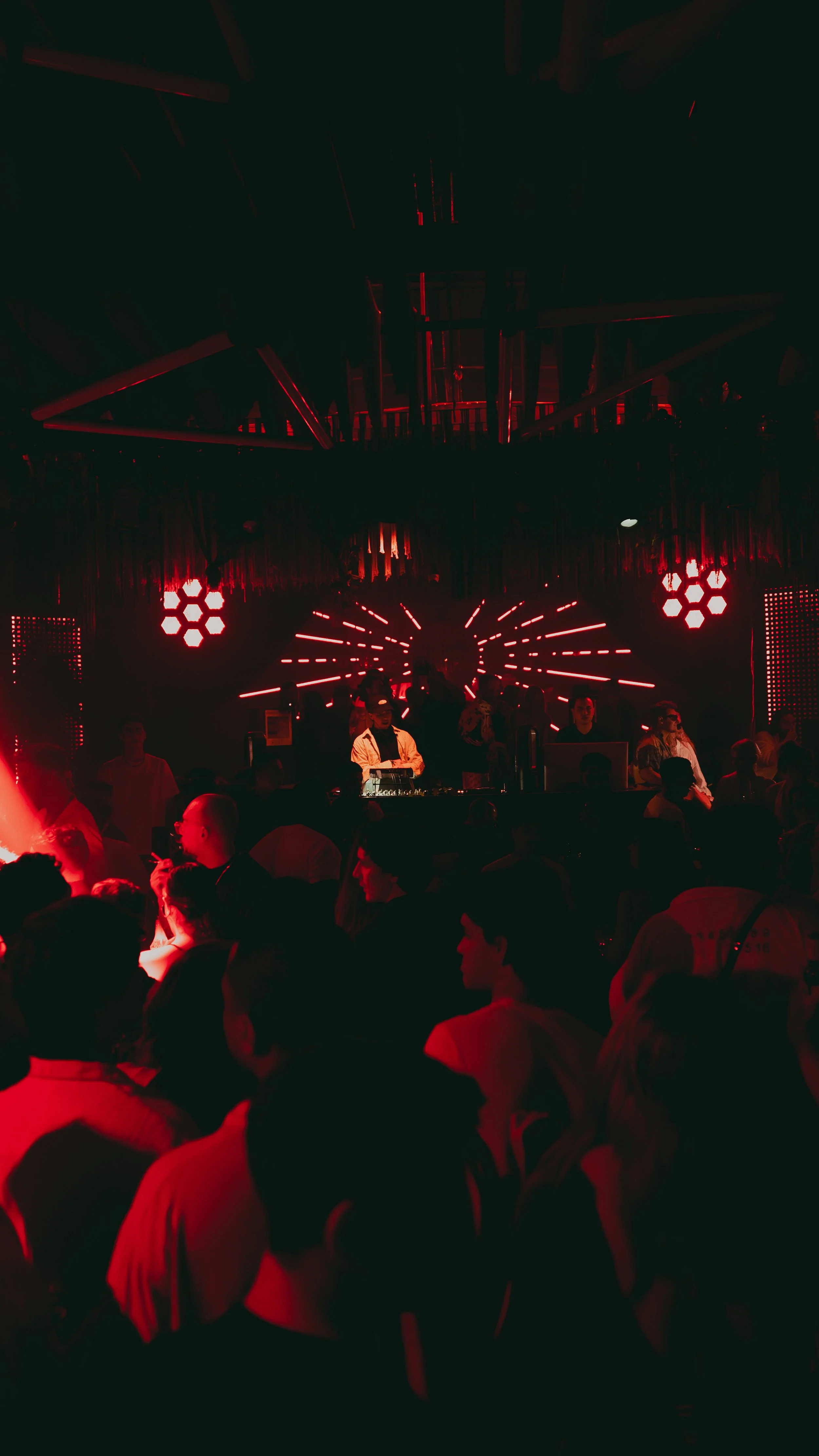 People dancing at a dimly lit nightclub with red lighting, a DJ playing music on a stage, and decorative LED light patterns overhead.