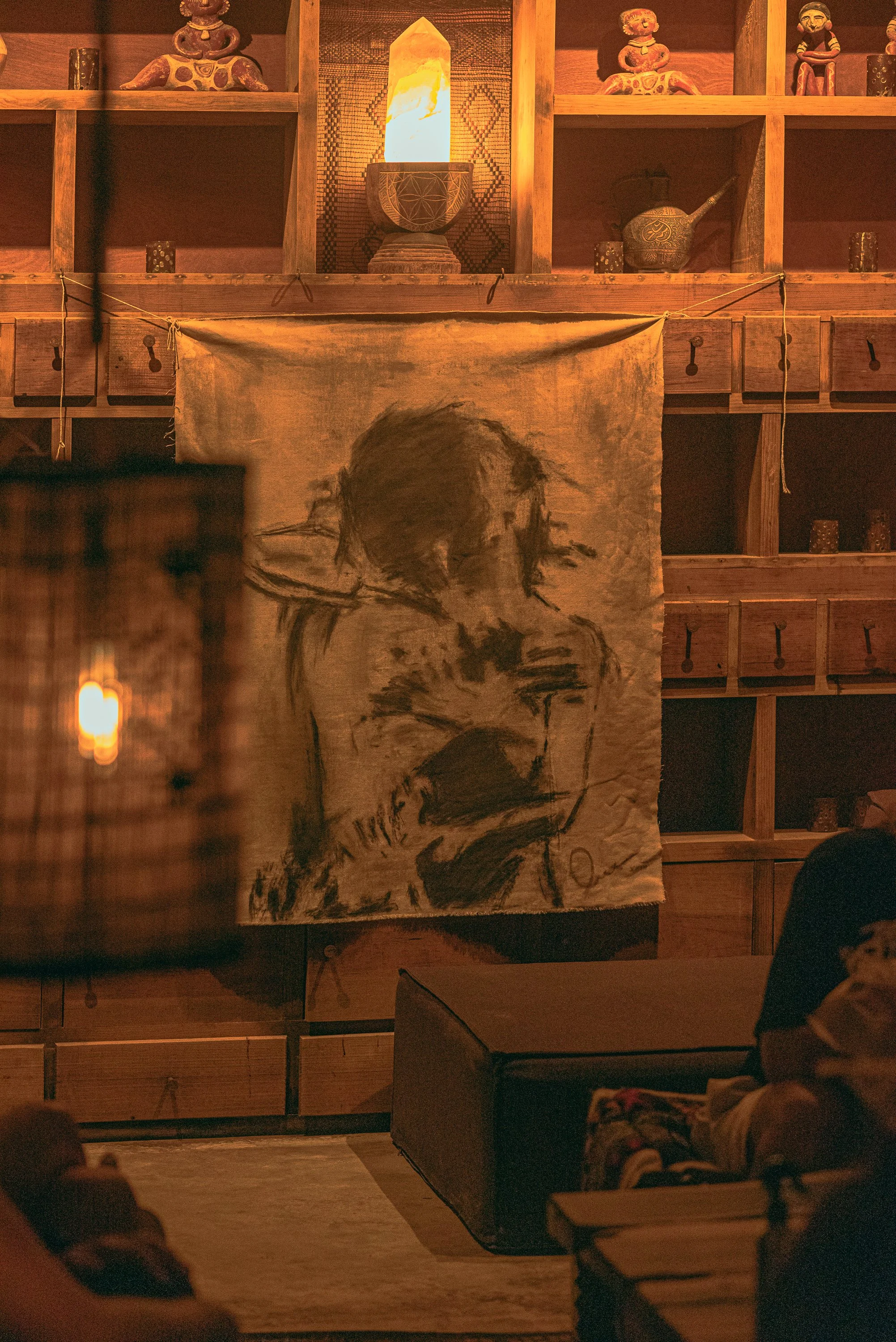 A cozy room with wooden shelves and decorative items, featuring a large, monochrome portrait of a person with long hair, holding their head in their hands, hanging on the wall.