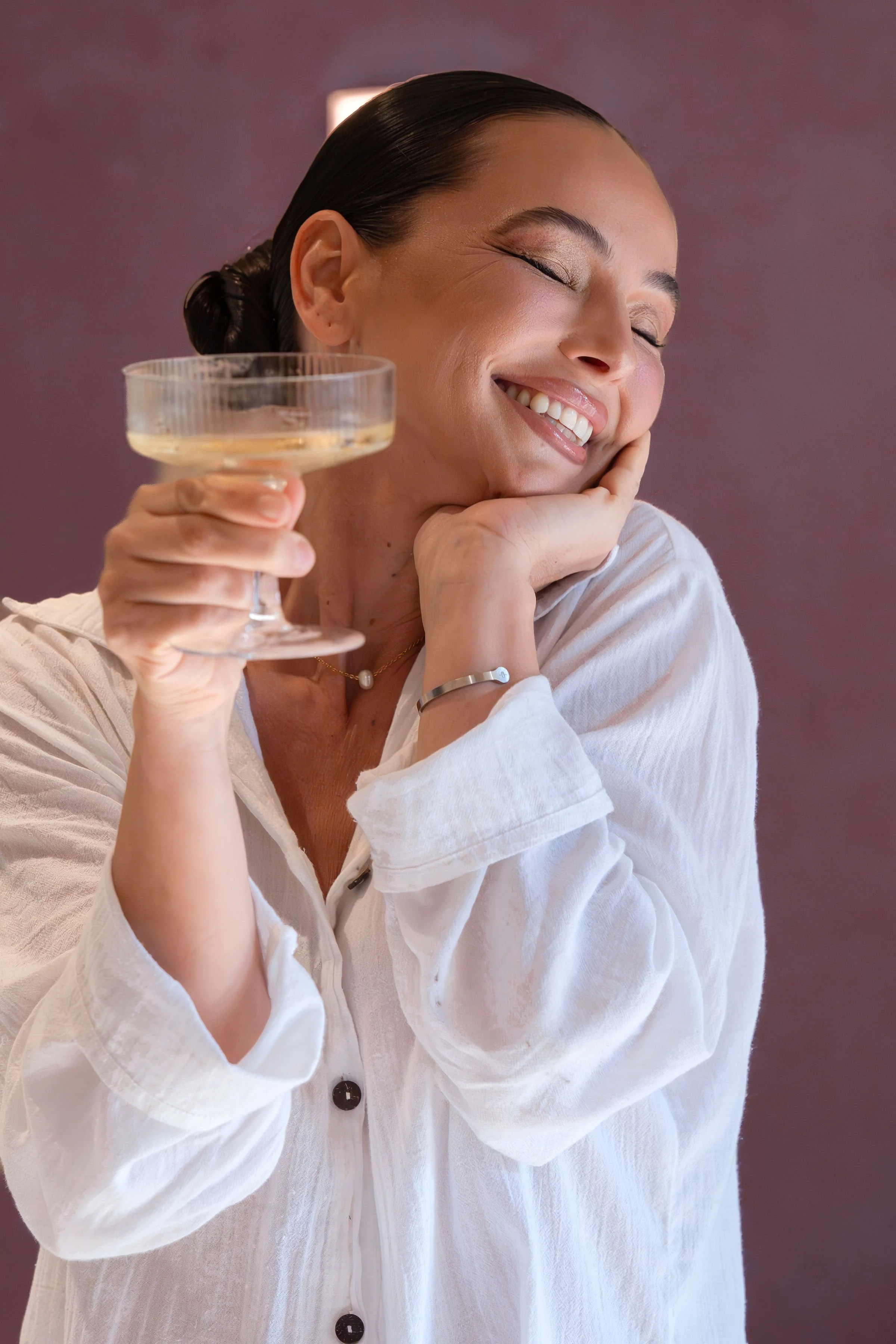 A woman smiling with eyes closed, holding a cocktail glass, dressed in a white shirt against a purple background.