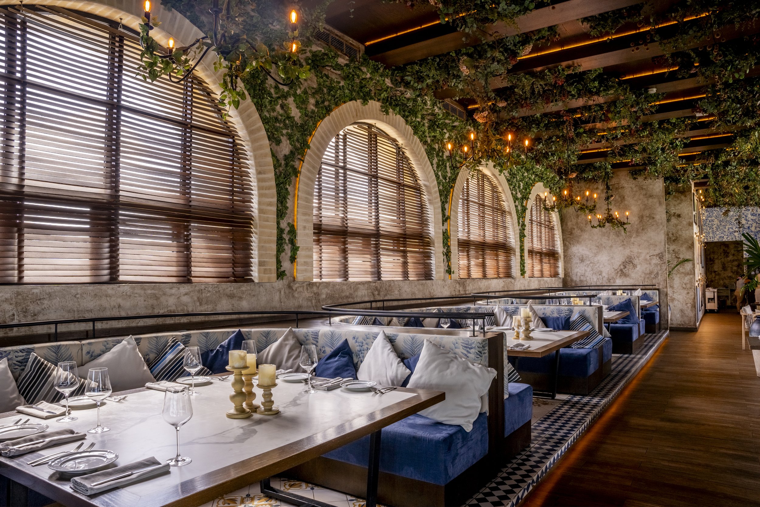 Interior of a restaurant with large arched windows, wooden blinds, greenery on the walls, chandeliers with candles, and blue cushioned booths with white and blue pillows, set with glassware and candles on the tables.