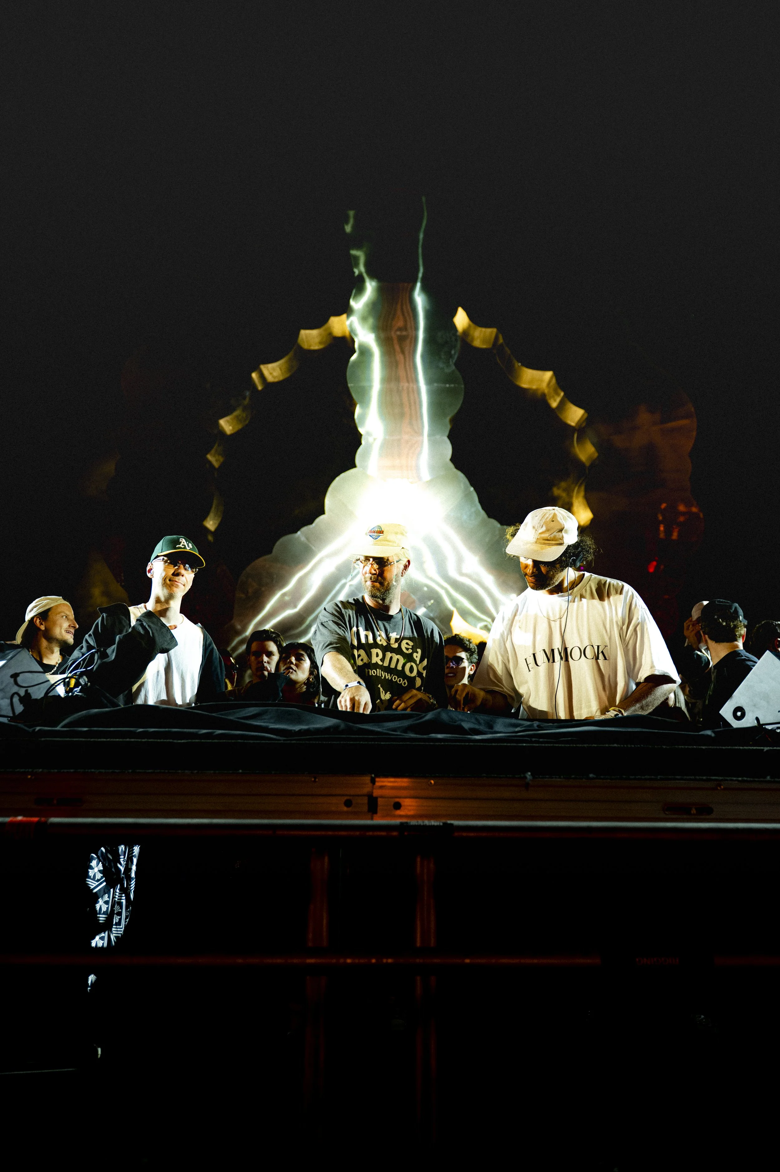 A group of DJs performing at a night event with a fiery and lightning visual display in the background.