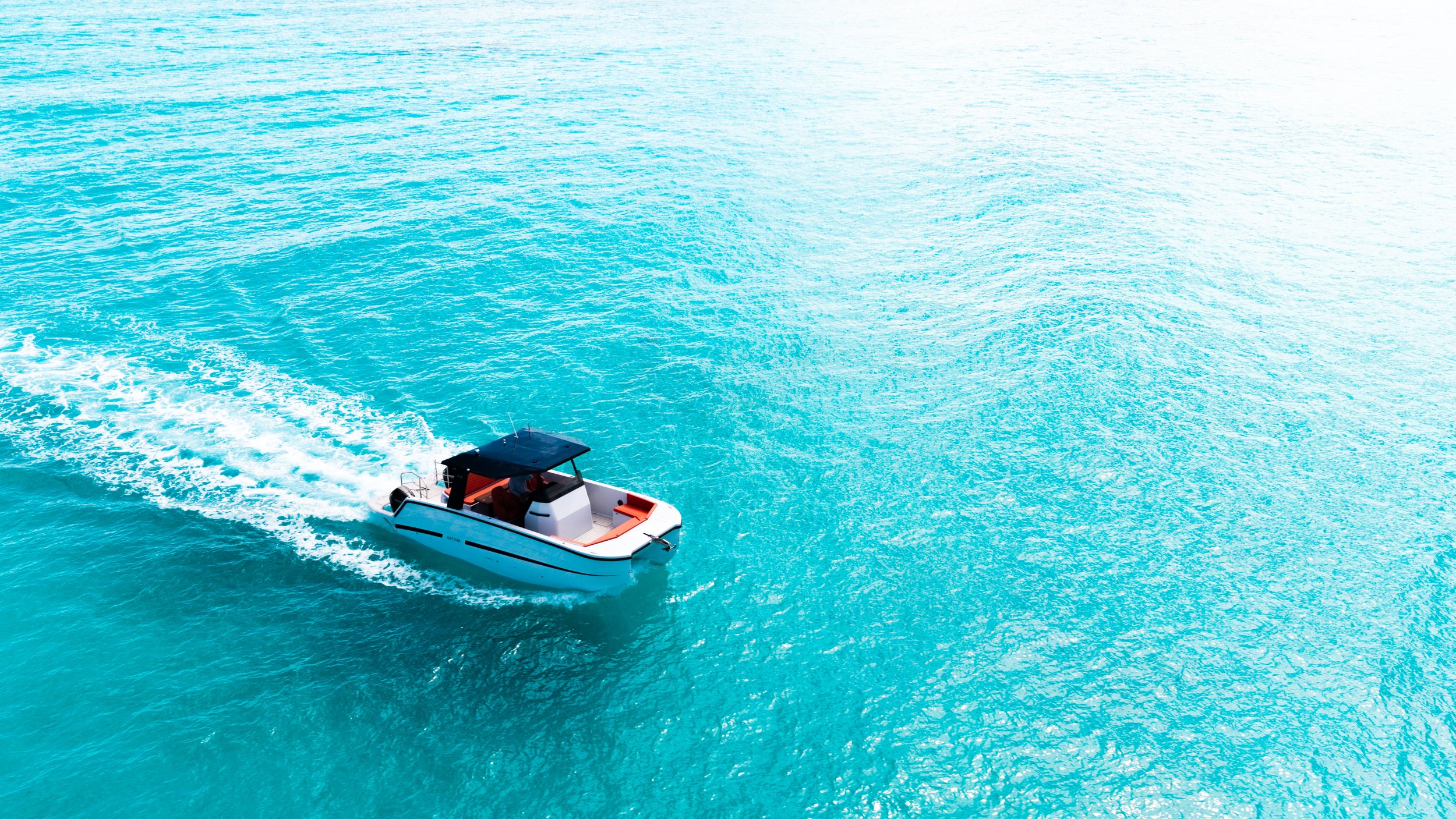 A small white boat with a black roof seen on the water, creating a wake as it moves through the calm, turquoise ocean.