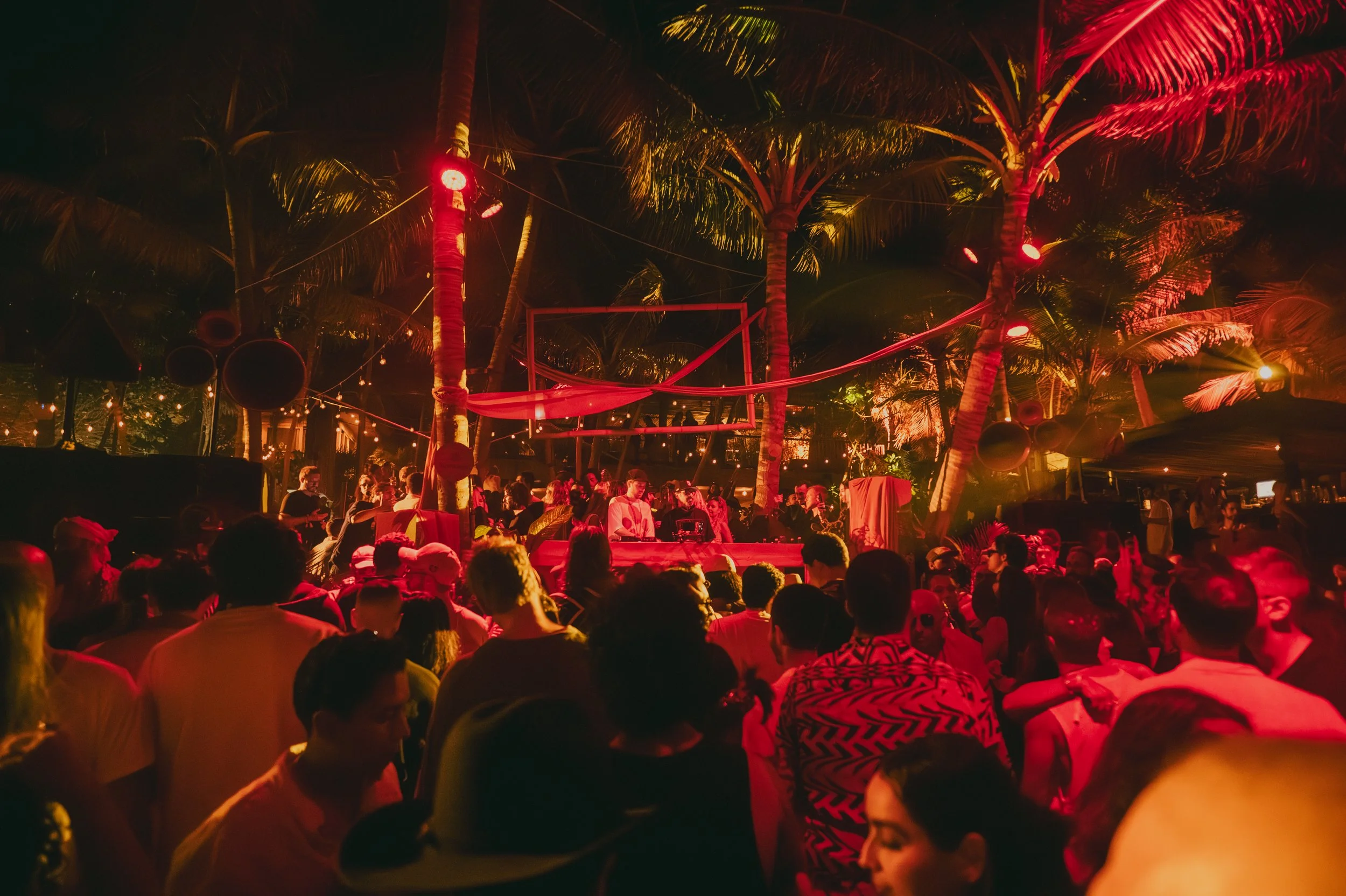 A lively nighttime outdoor party with a large crowd of people under palm trees illuminated by warm red and yellow lighting. There is a DJ or performer at a table in the center, with a makeshift basketball hoop and decorations hanging from trees.