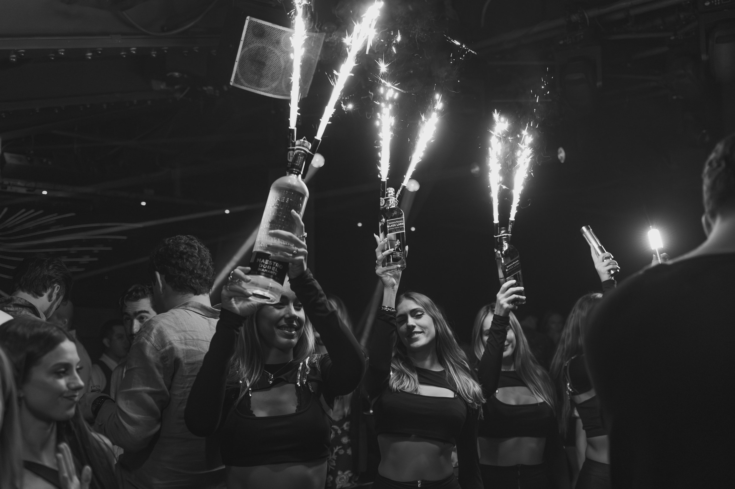 People celebrating at a party, holding bottles with sparklers attached, in a nightclub or party setting.