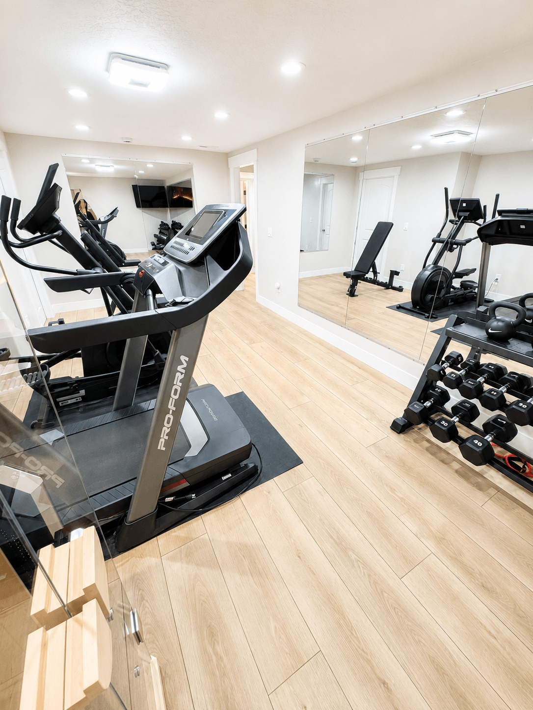 A home gym with a treadmill, weight bench, dumbbells, kettlebells, and cardio machines, with a large mirror on the wall.