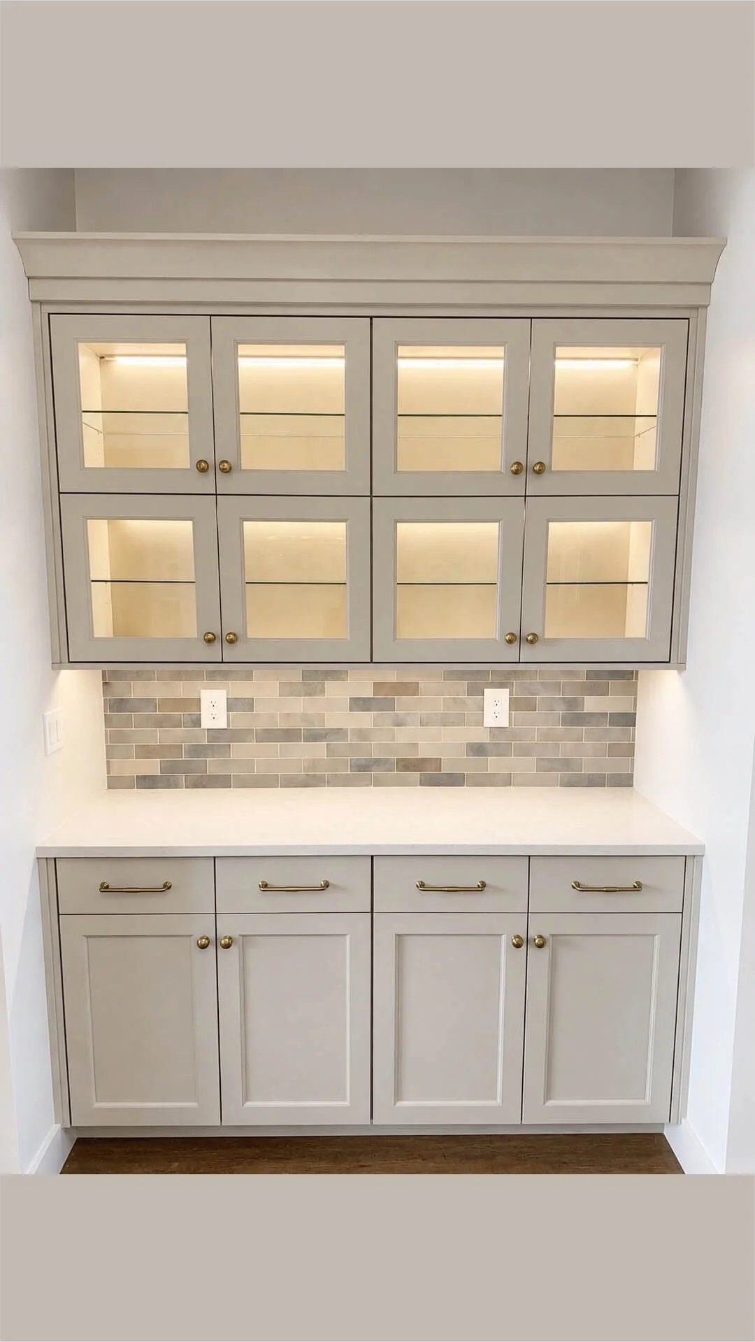 A built-in kitchen cabinet with glass-paneled upper doors, illuminated inside, and lower cabinets with gold handles, set against a gray brick backsplash and white countertop.