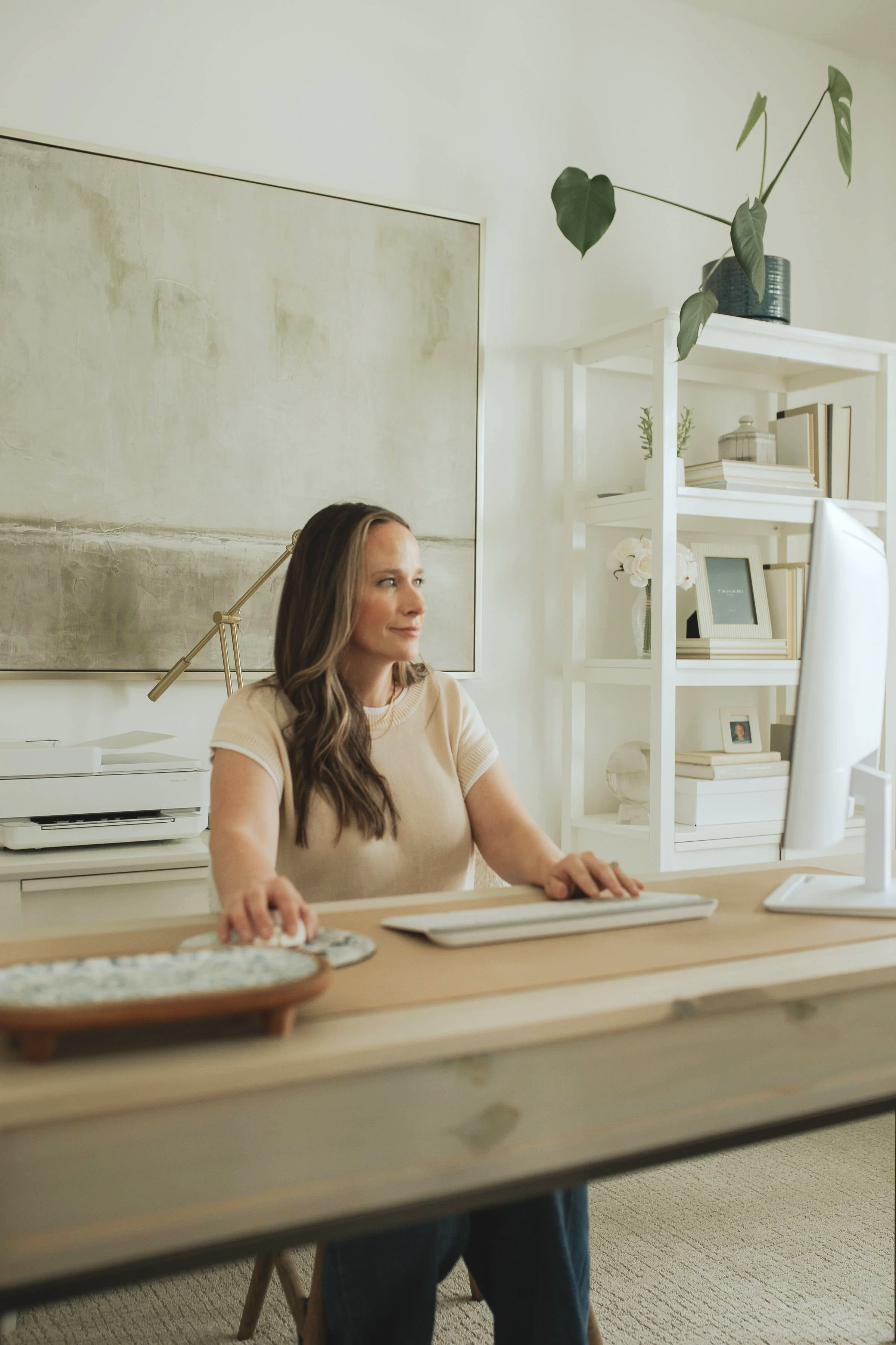 A woman sitting at a desk working on a computer in a bright, minimalist home office with white shelves and decorative items.
