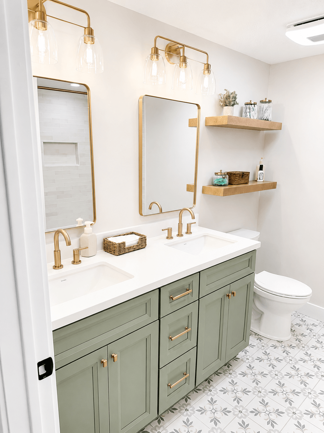 A modern bathroom with double sink vanity, green cabinets with gold handles, two rectangular mirrors with gold frames, and warm lighting fixtures. There are wooden floating shelves with decorative items, a toilet, and patterned tile flooring.
