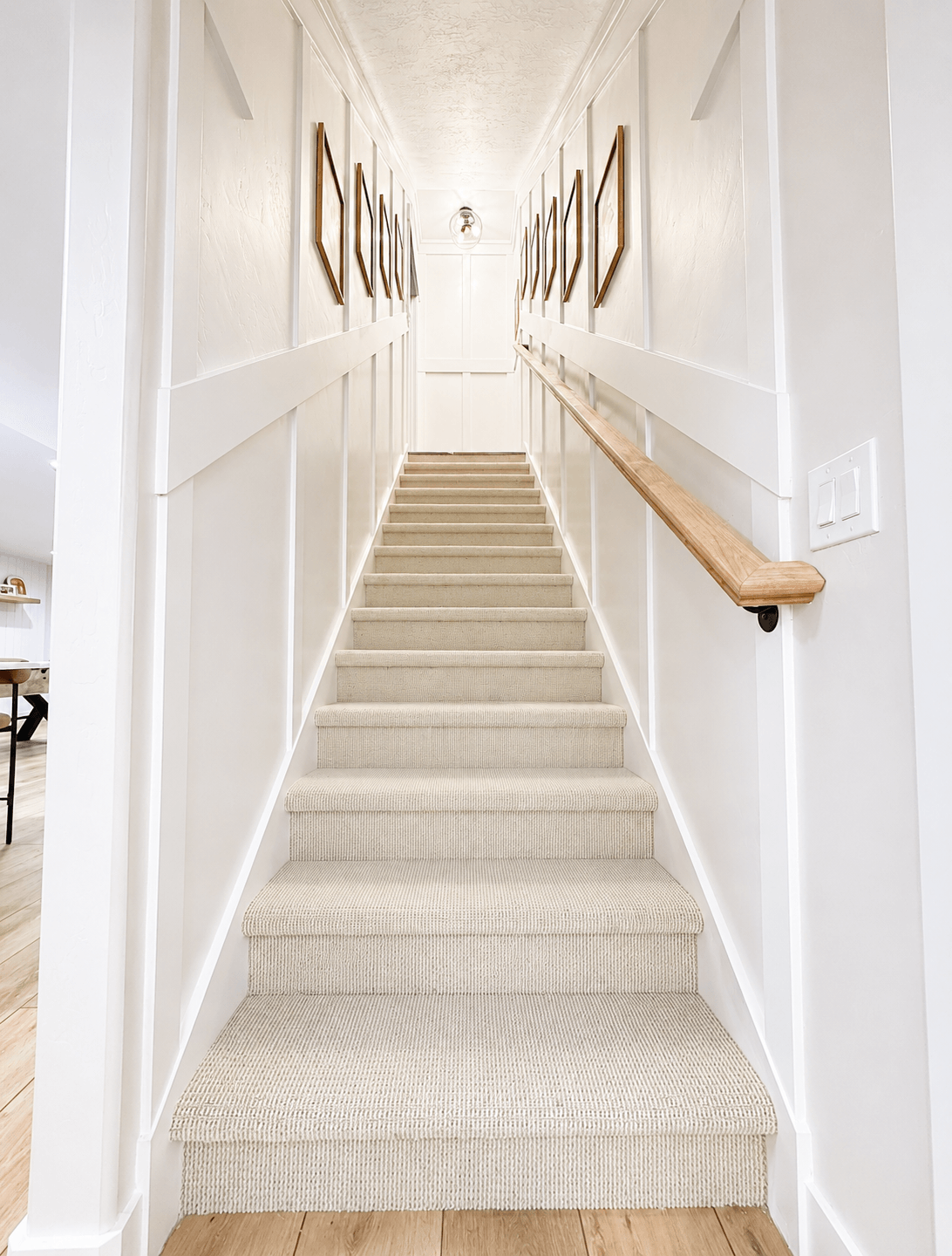 Interior view of a staircase with beige carpeting, white walls, framed artwork on both sides, a wooden handrail on the right, and ceiling light fixtures at the top.