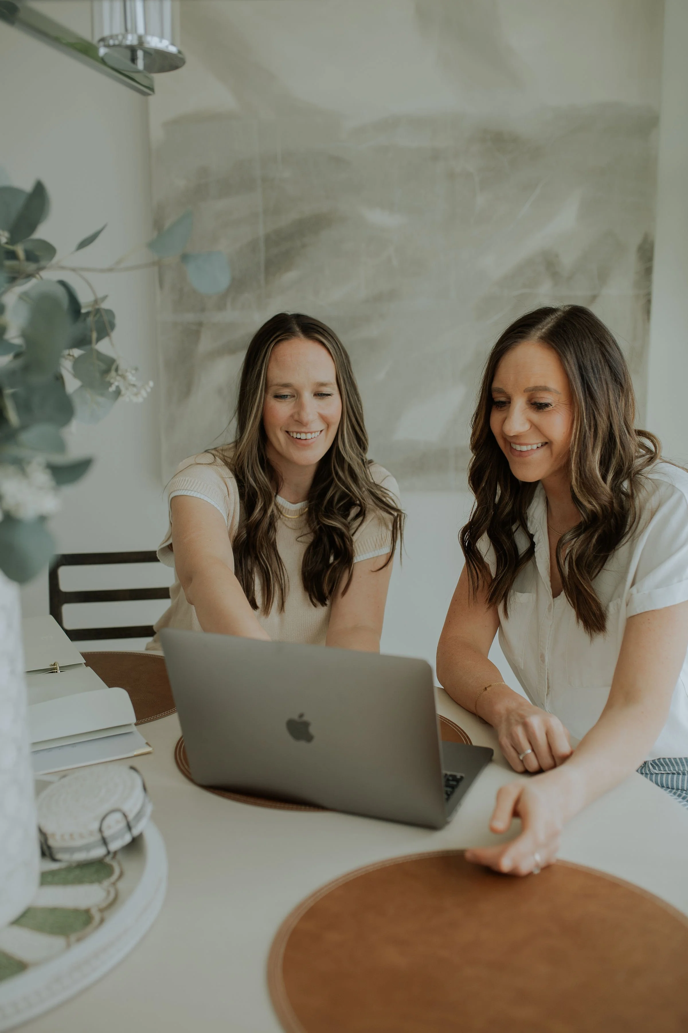 Two women sitting at a table working on a laptop in a well-lit room with a decorative backsplash and a plant in the foreground.