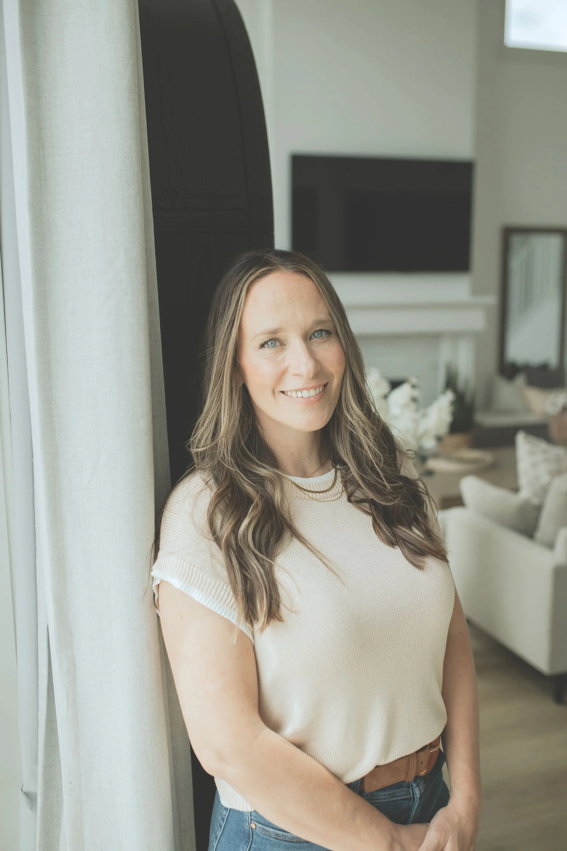 A woman with long wavy brown hair and blue eyes smiling, standing next to a window in a cozy, modern living room.